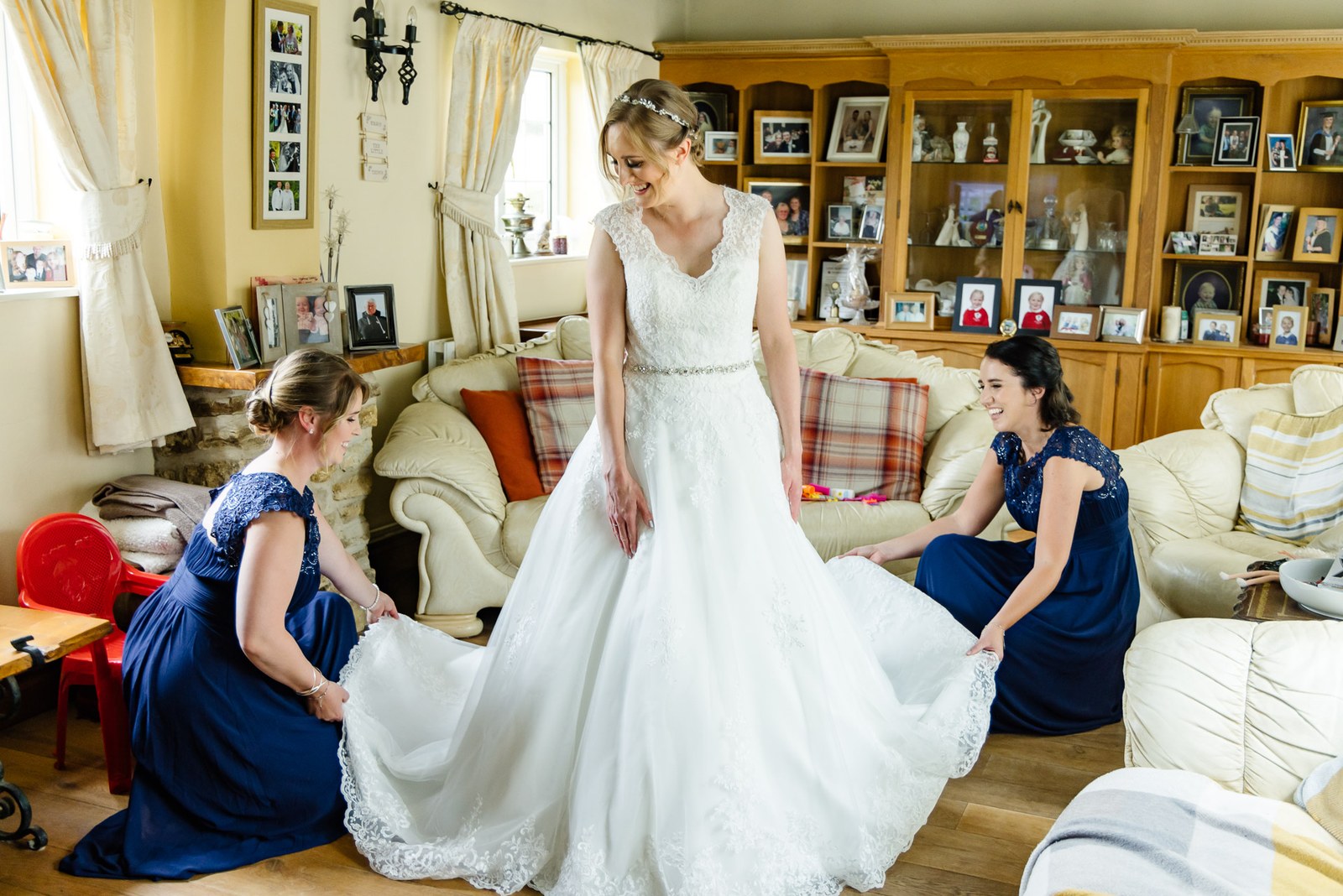 Bridesmaids in navy help a bride into her gown in a cottage sitting room