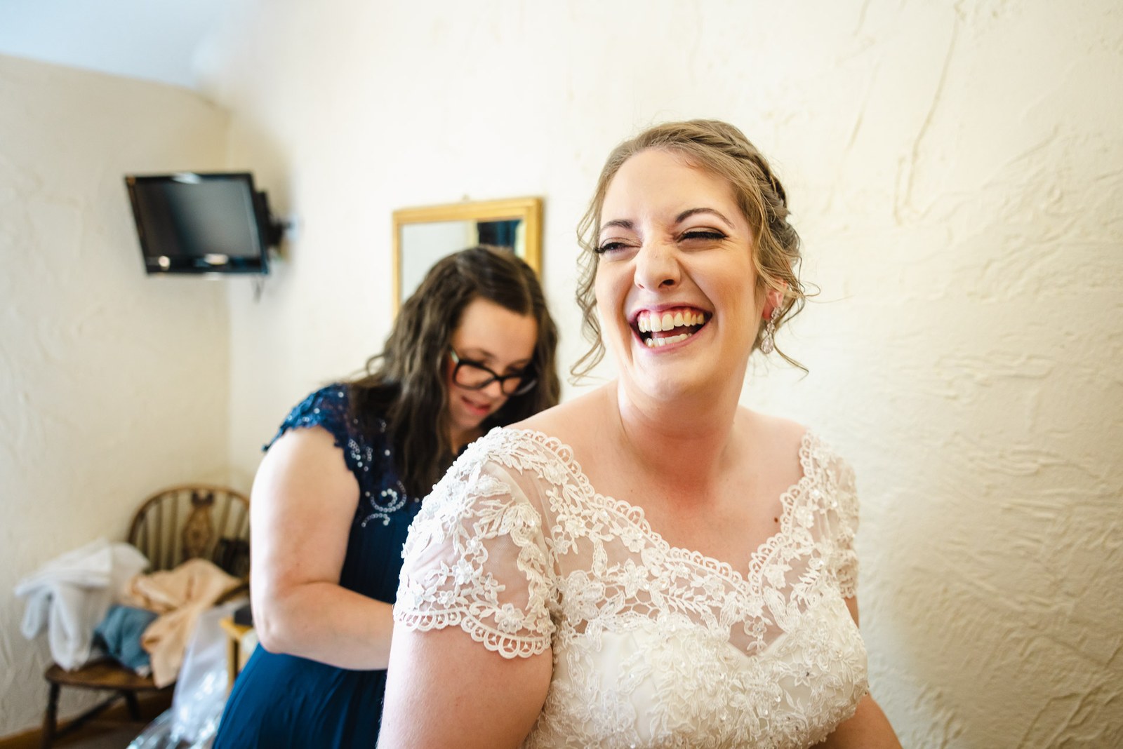 Bride laughing as a bridesmaid helps her dress against a pale cottage wall