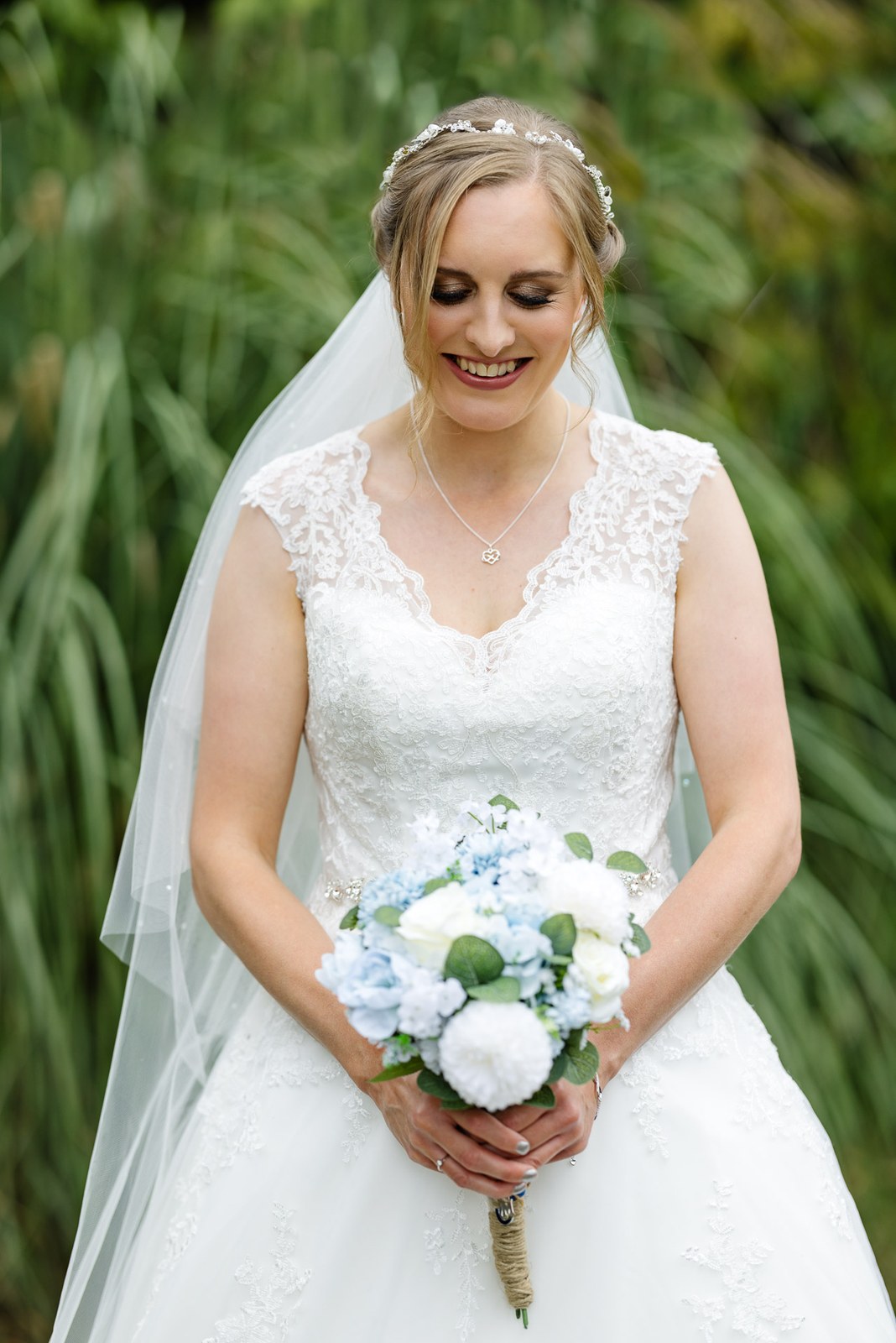Bride in a lace gown and veil holding a blue and white bouquet by tall grasses