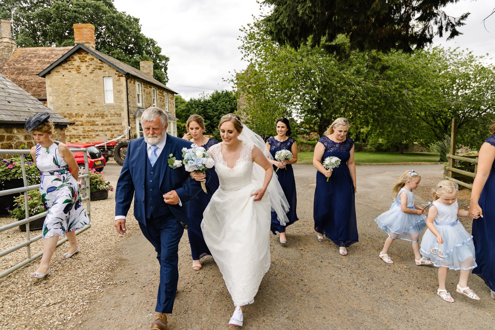 Father walks a bride along the gravel drive at Slapton Manor with bridesmaids following