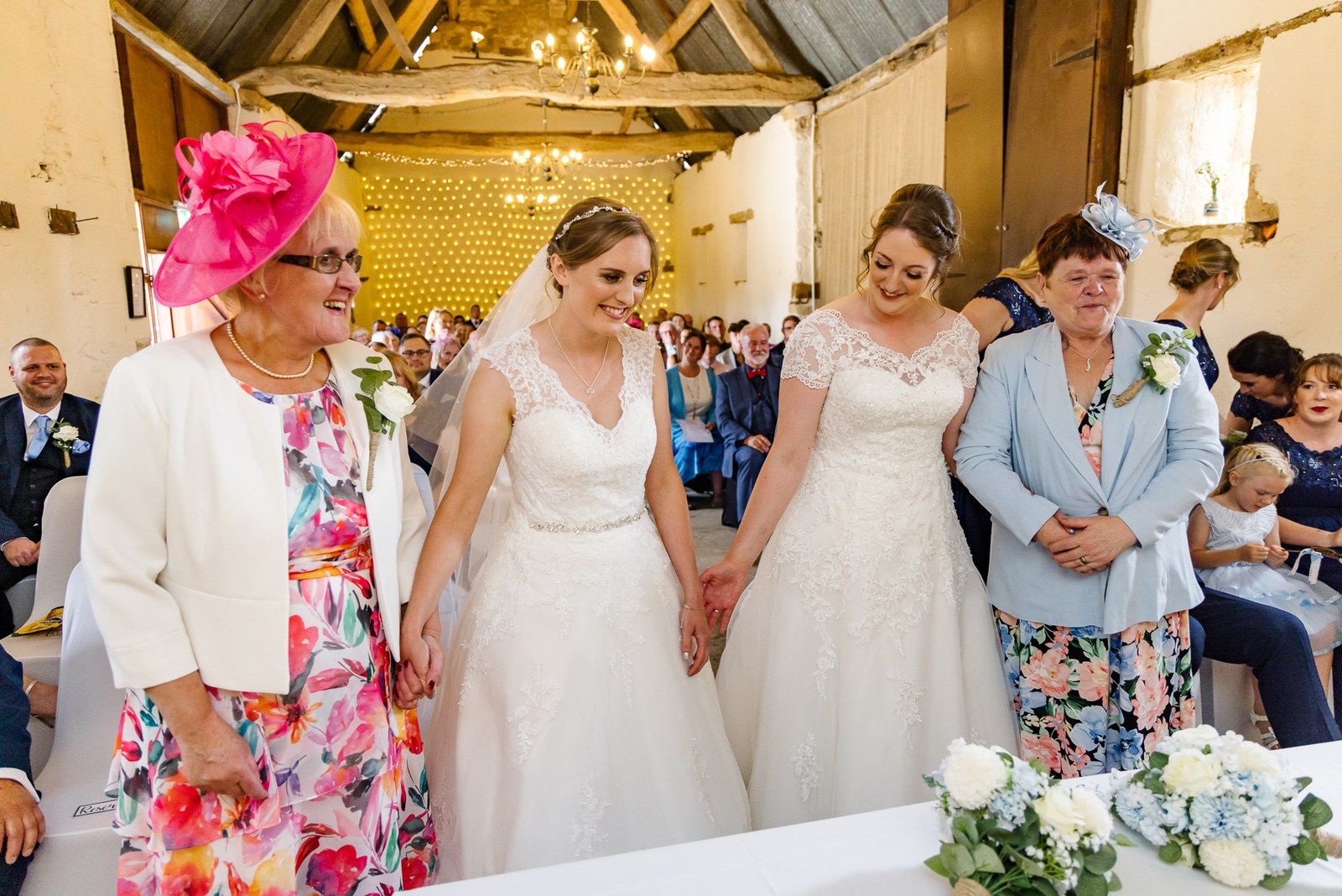 The two brides clasp hands at the ceremony as their mothers look on