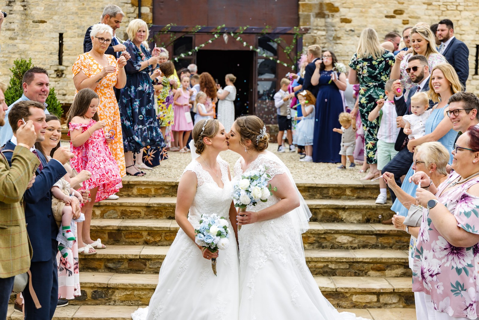 The two brides kiss on the manor's stone steps as guests blow bubbles
