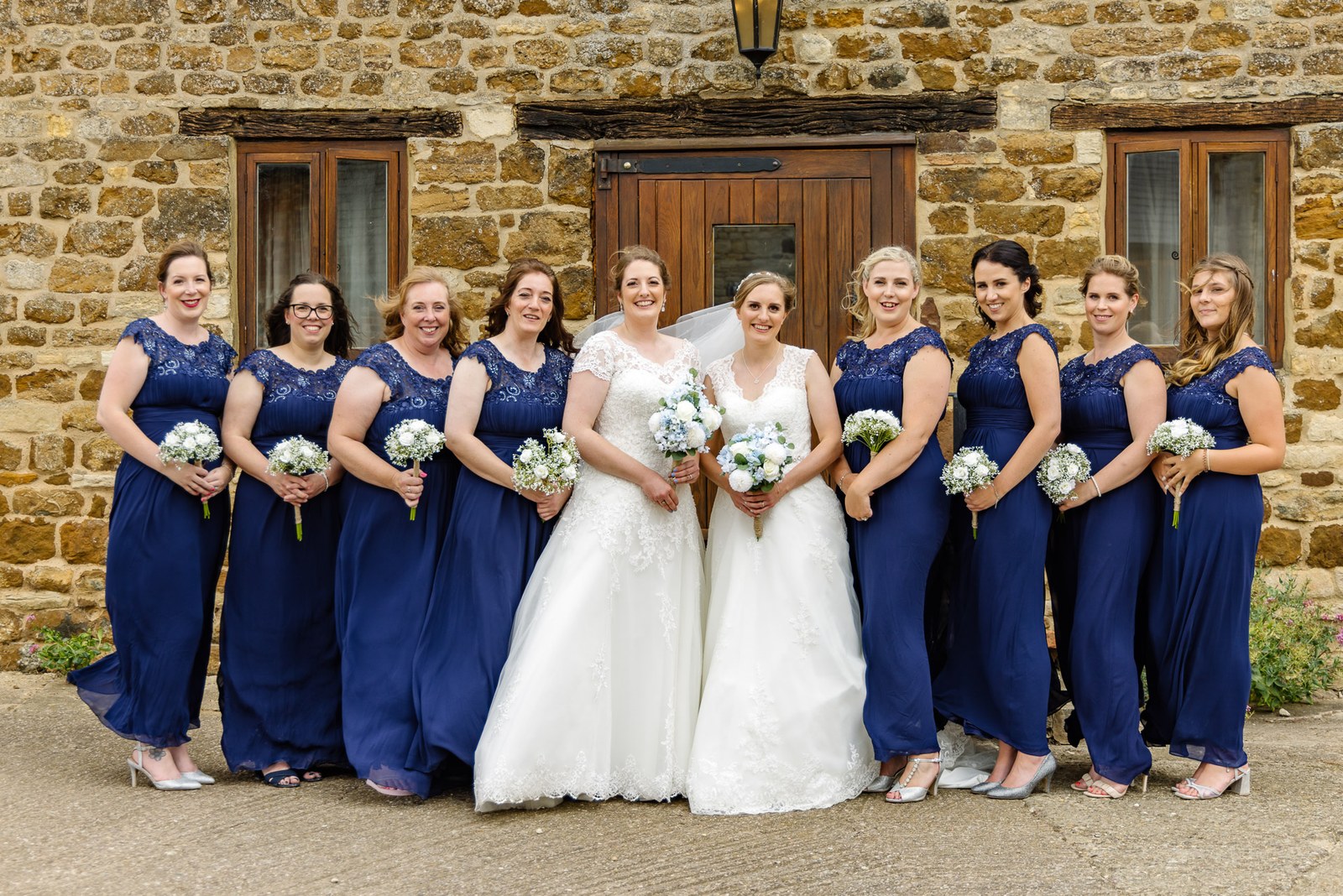 The two brides with their bridesmaids in navy by the stone barn