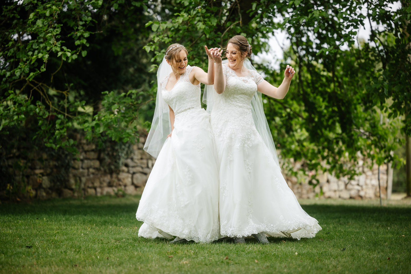 The two brides hold hands beneath leafy trees on the Slapton Manor lawn