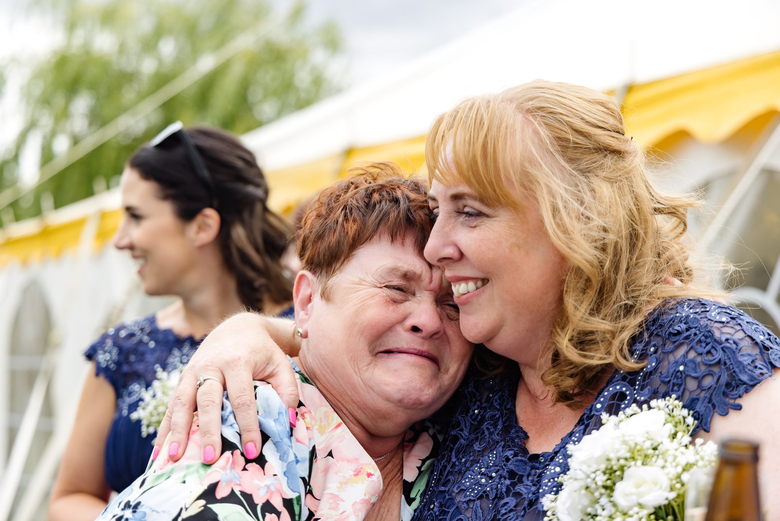 A bridesmaid in navy hugs a tearful older guest outside the marquee