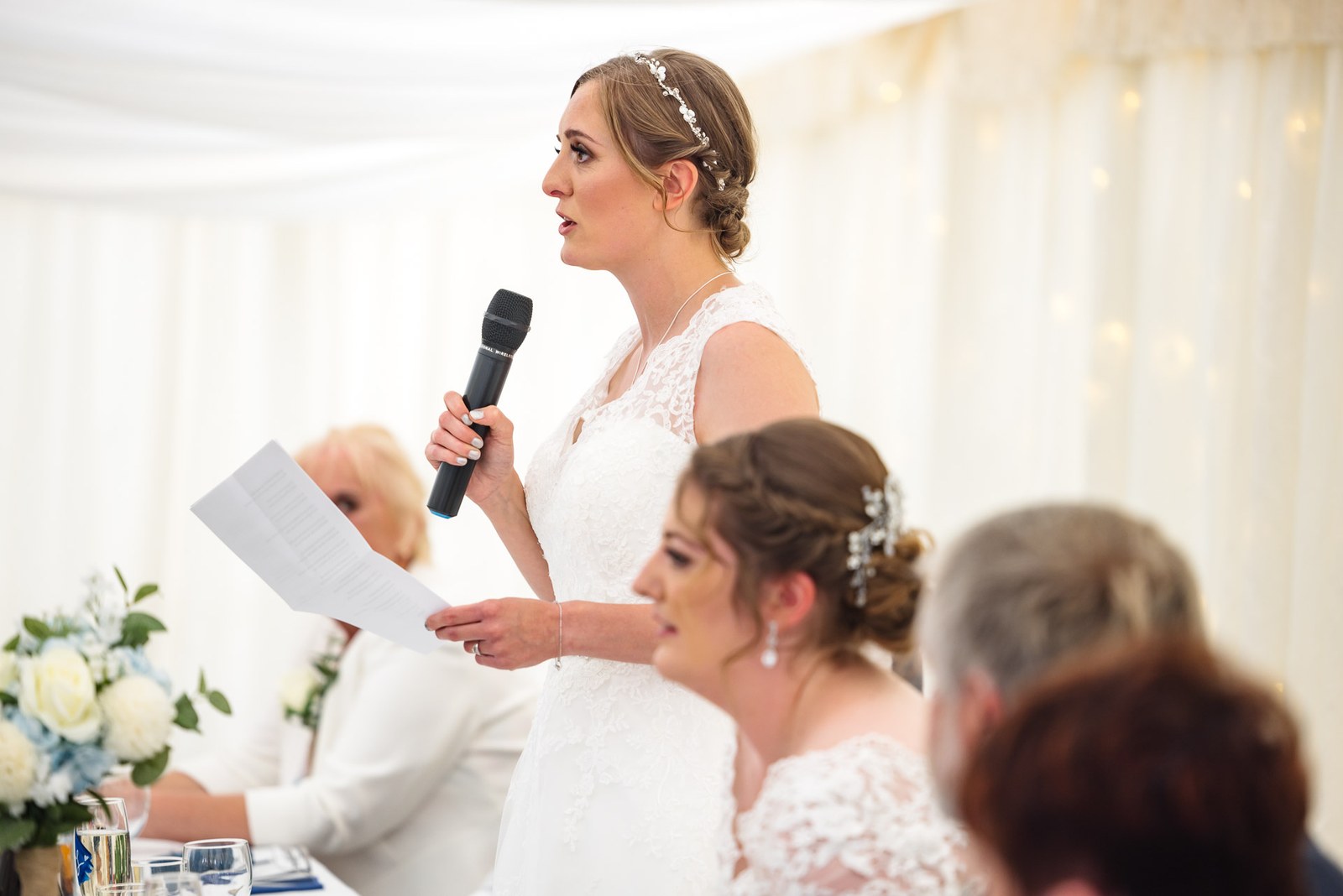 A bride delivers a speech into a microphone inside the marquee