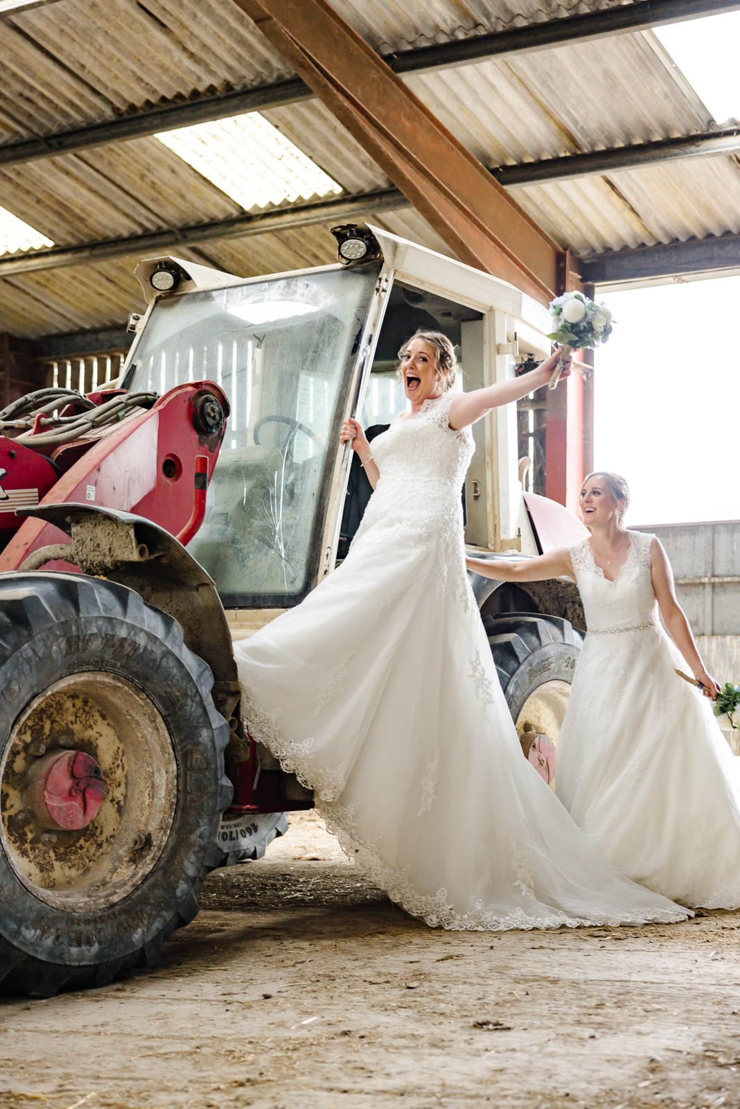 One bride laughs holding her bouquet aloft beside a red farm tractor