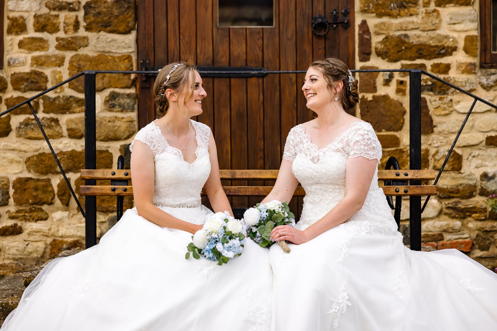 The two brides sit on a bench before the timber barn door at Slapton Manor