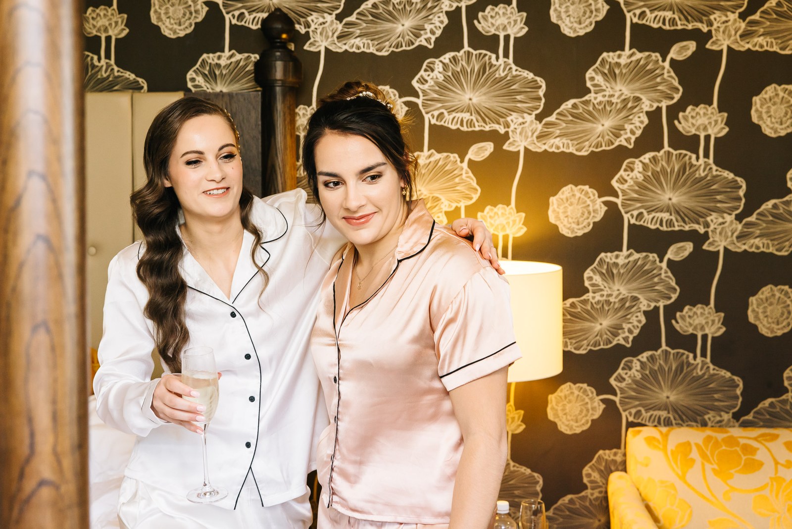 Bride and a bridesmaid in satin pyjamas raise a glass against floral wallpaper