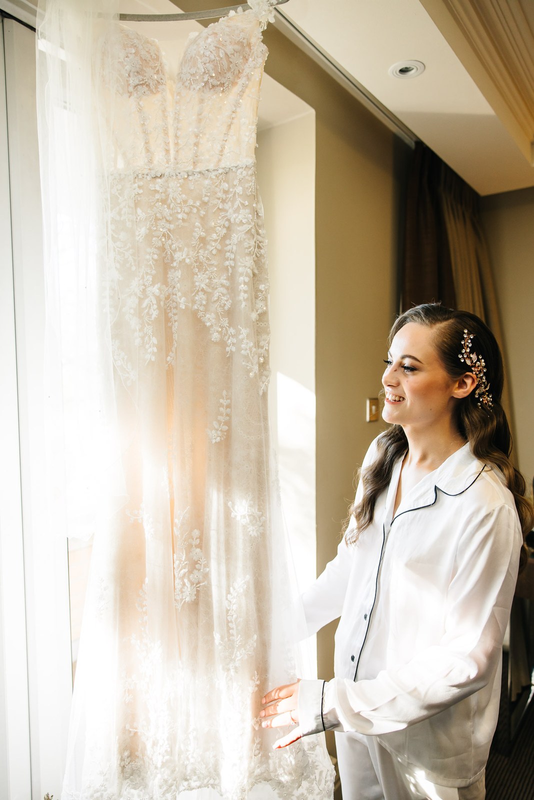 Bride in pyjamas by the window, her gown glowing in the light beside her