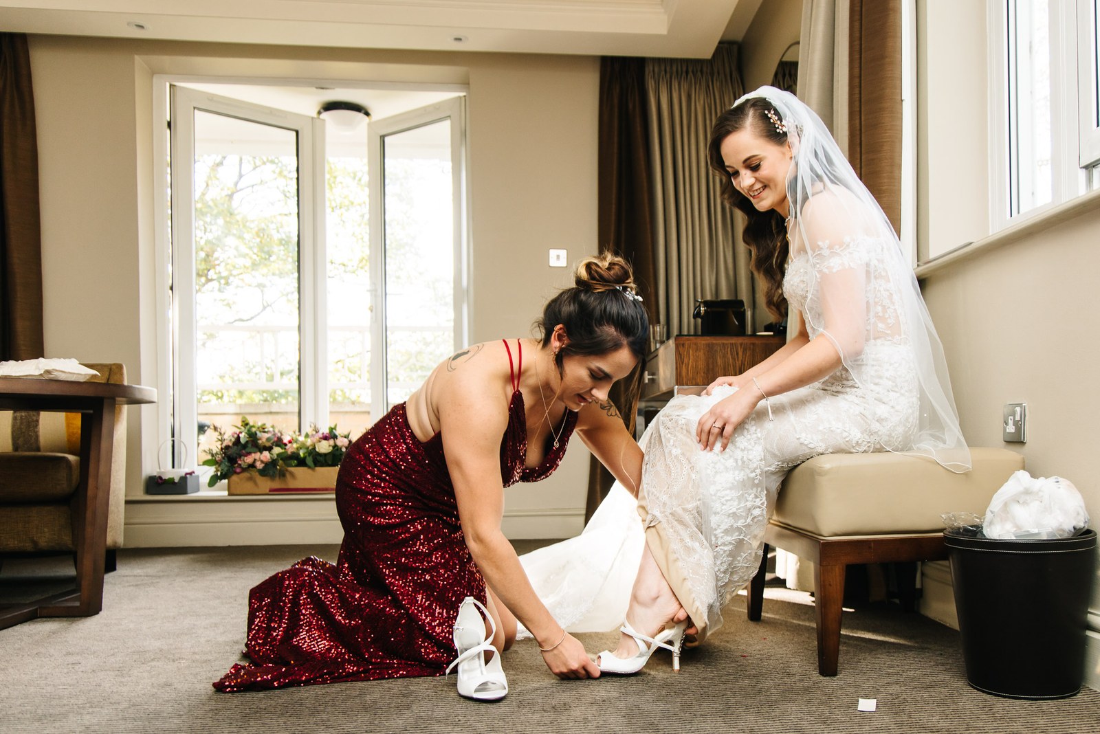 A bridesmaid in red helps the bride into her shoe by the window
