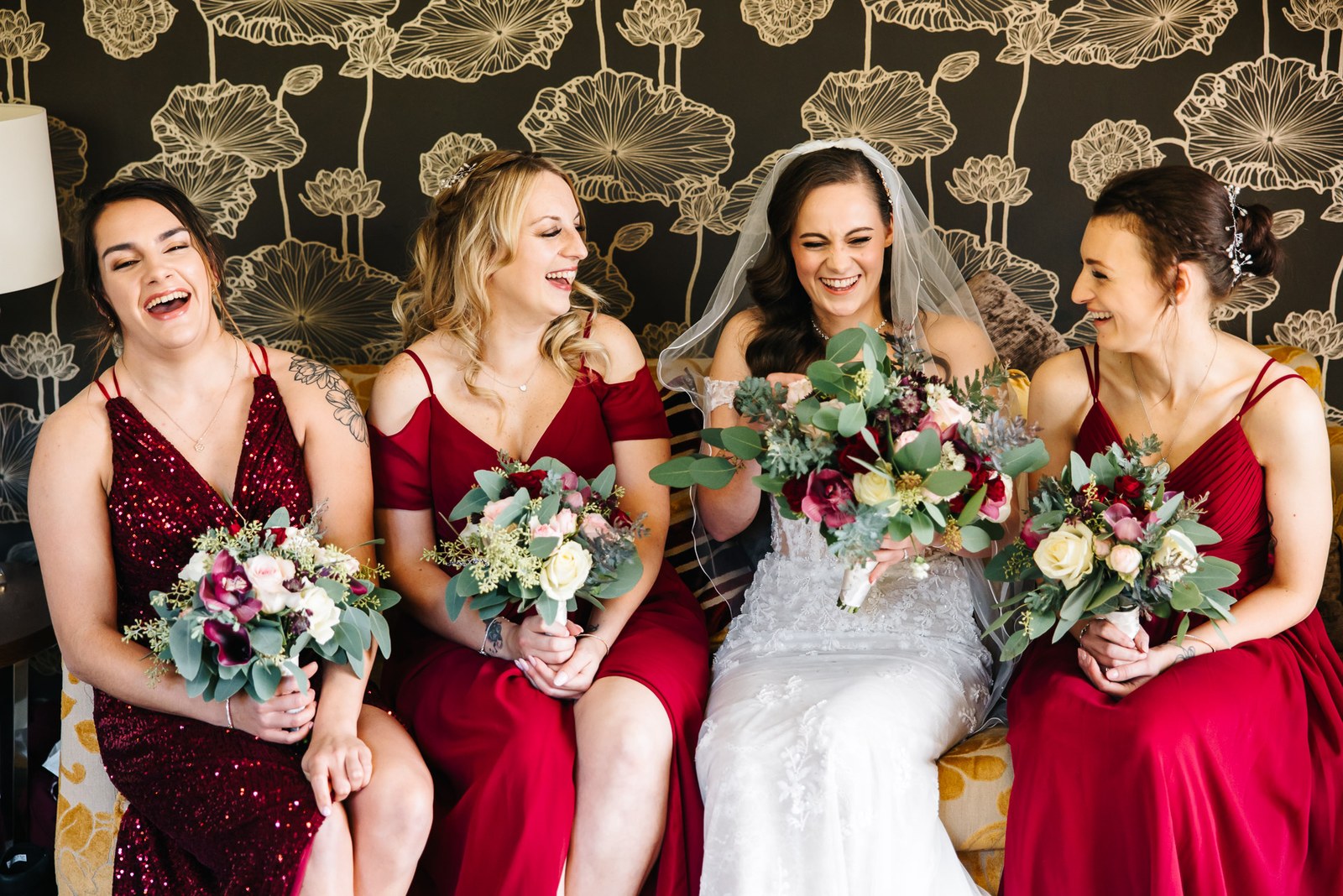 Bride seated with her red-gowned bridesmaids, all holding rich seasonal bouquets