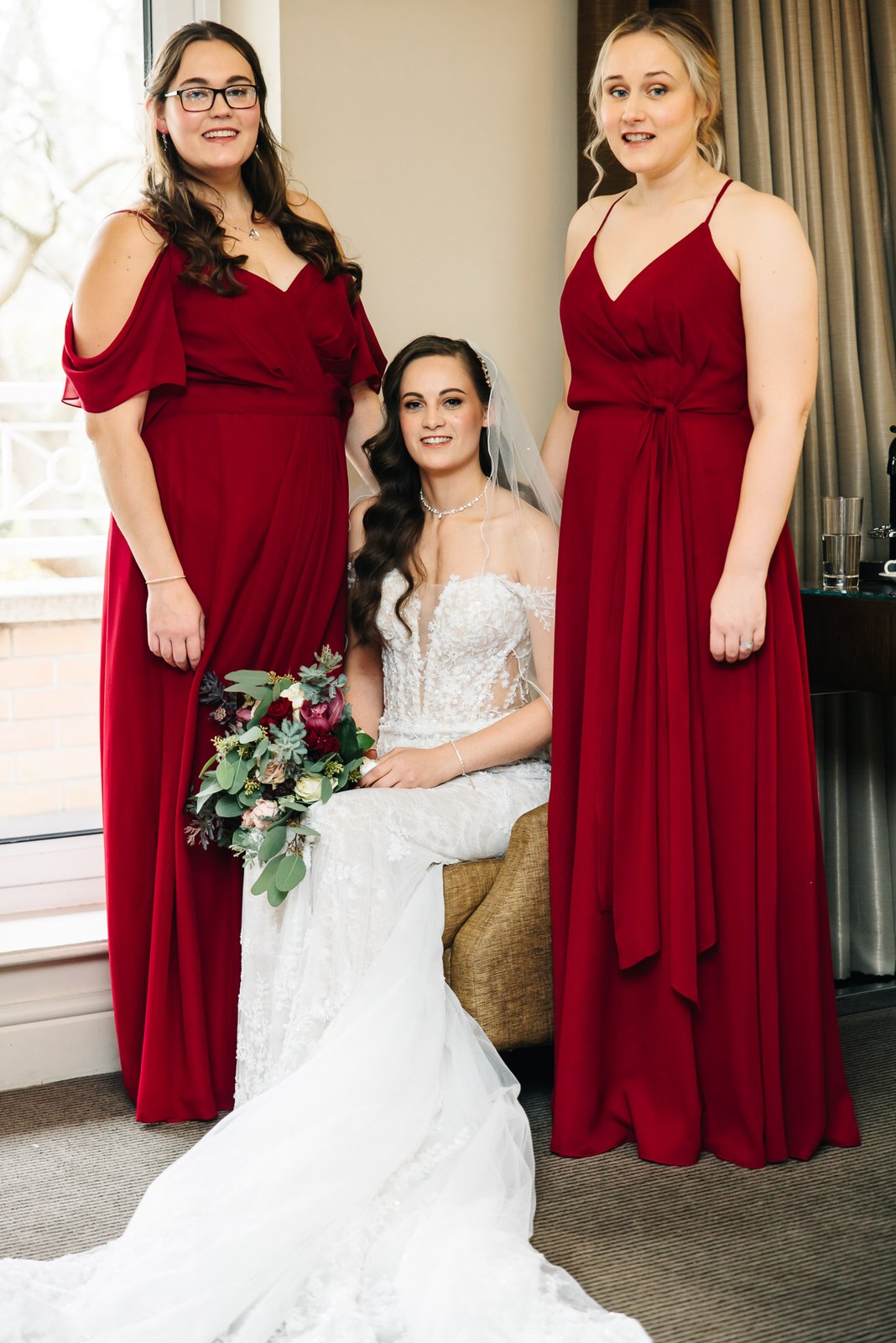 The bride flanked by two bridesmaids in deep red by the window