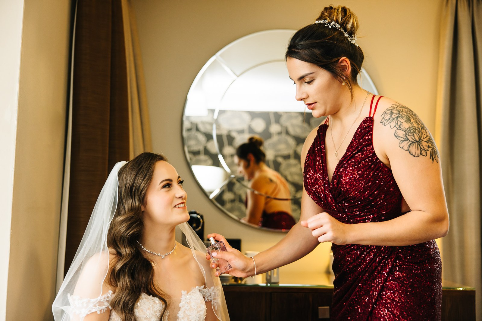 A bridesmaid in red spritzes the bride with perfume before a round mirror