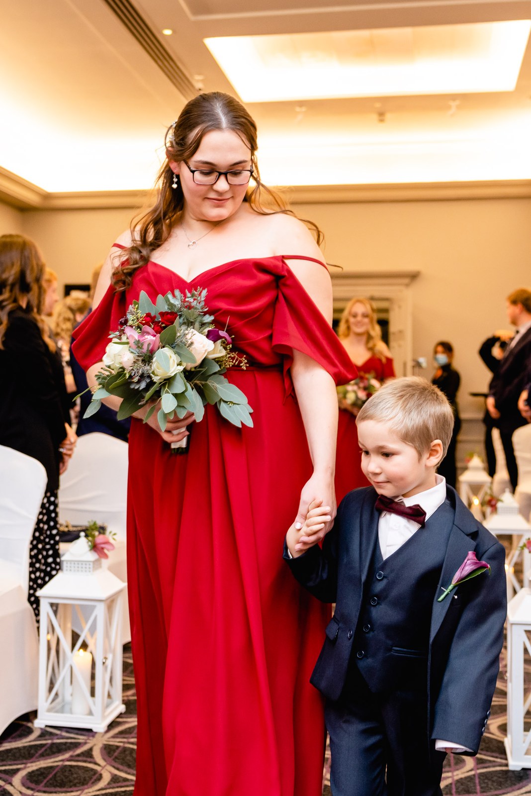A bridesmaid in red leads a page boy by the hand down the aisle