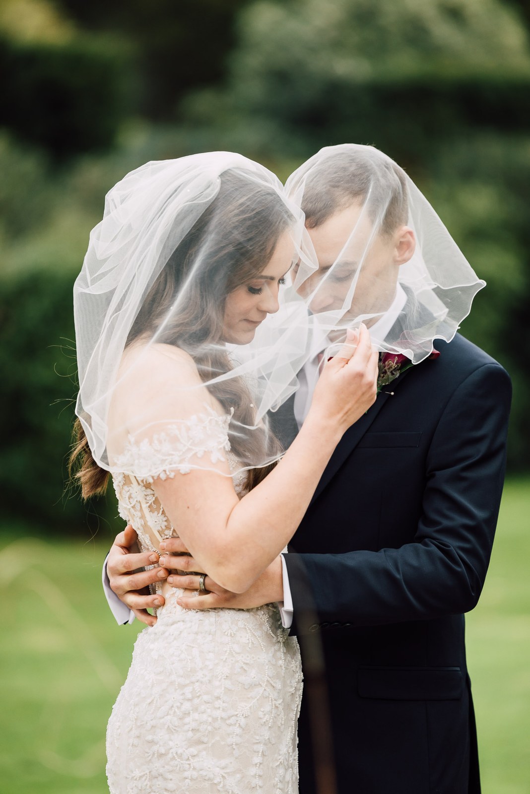 The couple embrace wrapped beneath the bride's veil in the grounds