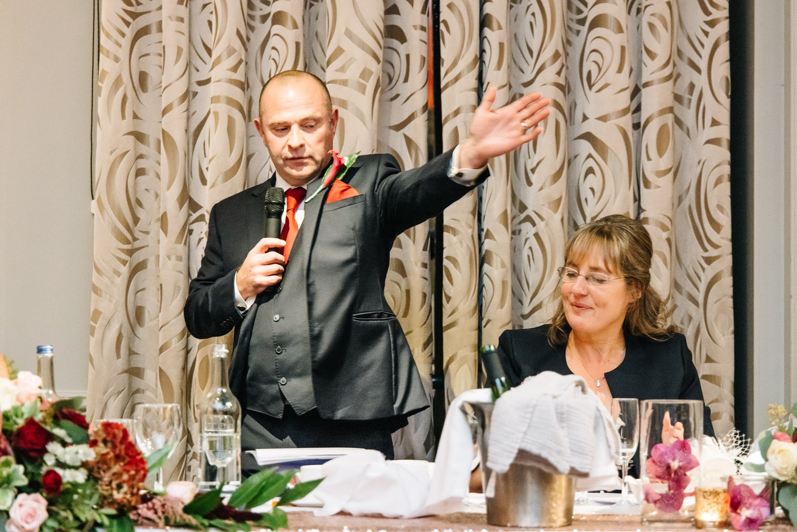 A speaker at the top table mid-speech with an arm thrown wide, mother of the bride beside him