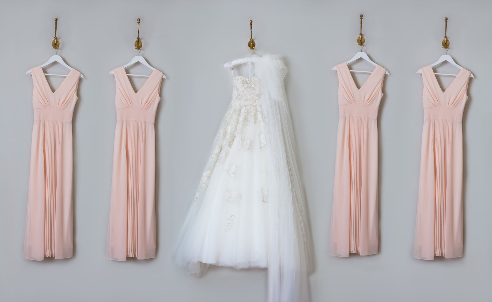 Four blush bridesmaid dresses hung beside the white wedding gown on a wall