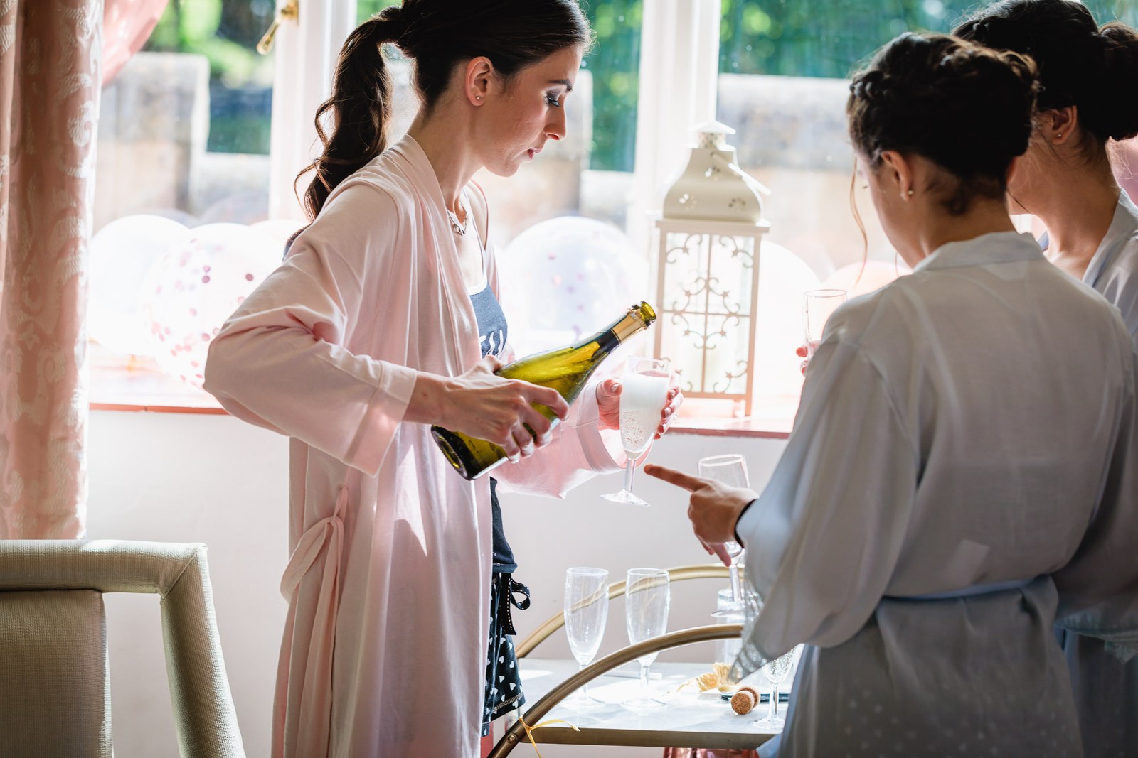 A bridesmaid in a robe pours fizz as the others get ready by the window