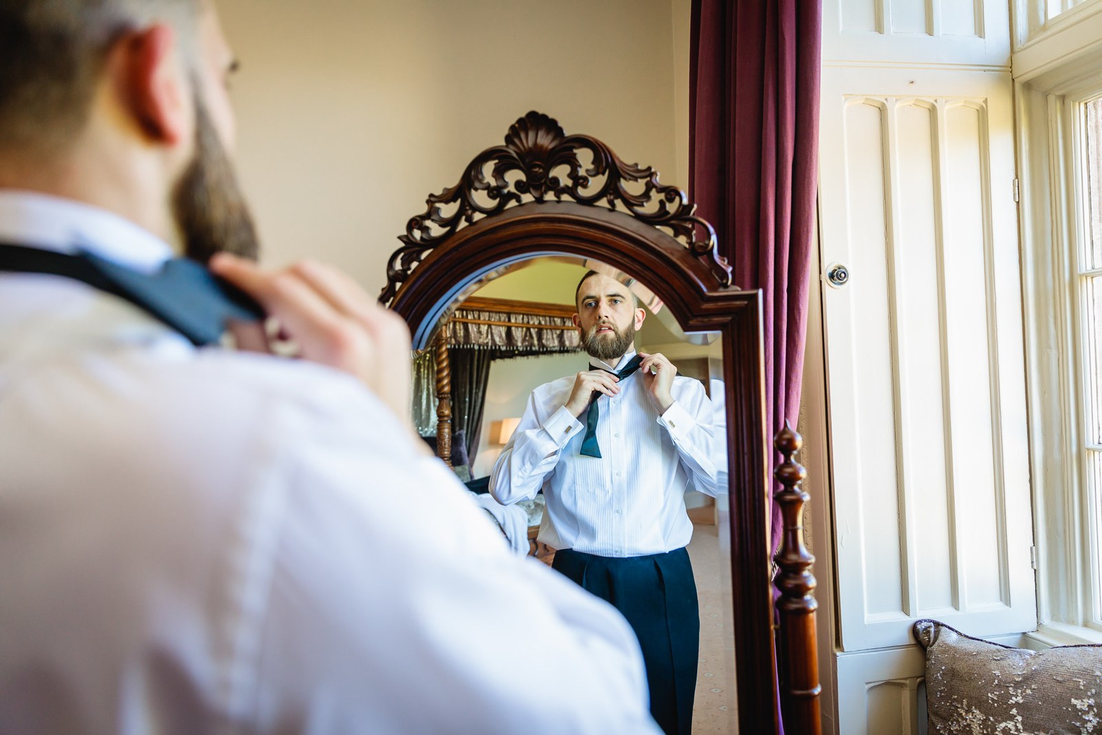 A man in shirtsleeves knots his tie before an ornate carved mirror