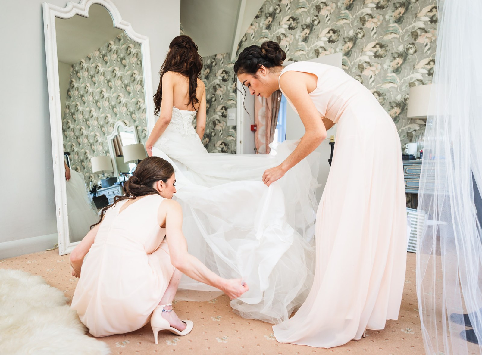 Bridesmaids help the bride into her gown before a tall mirror