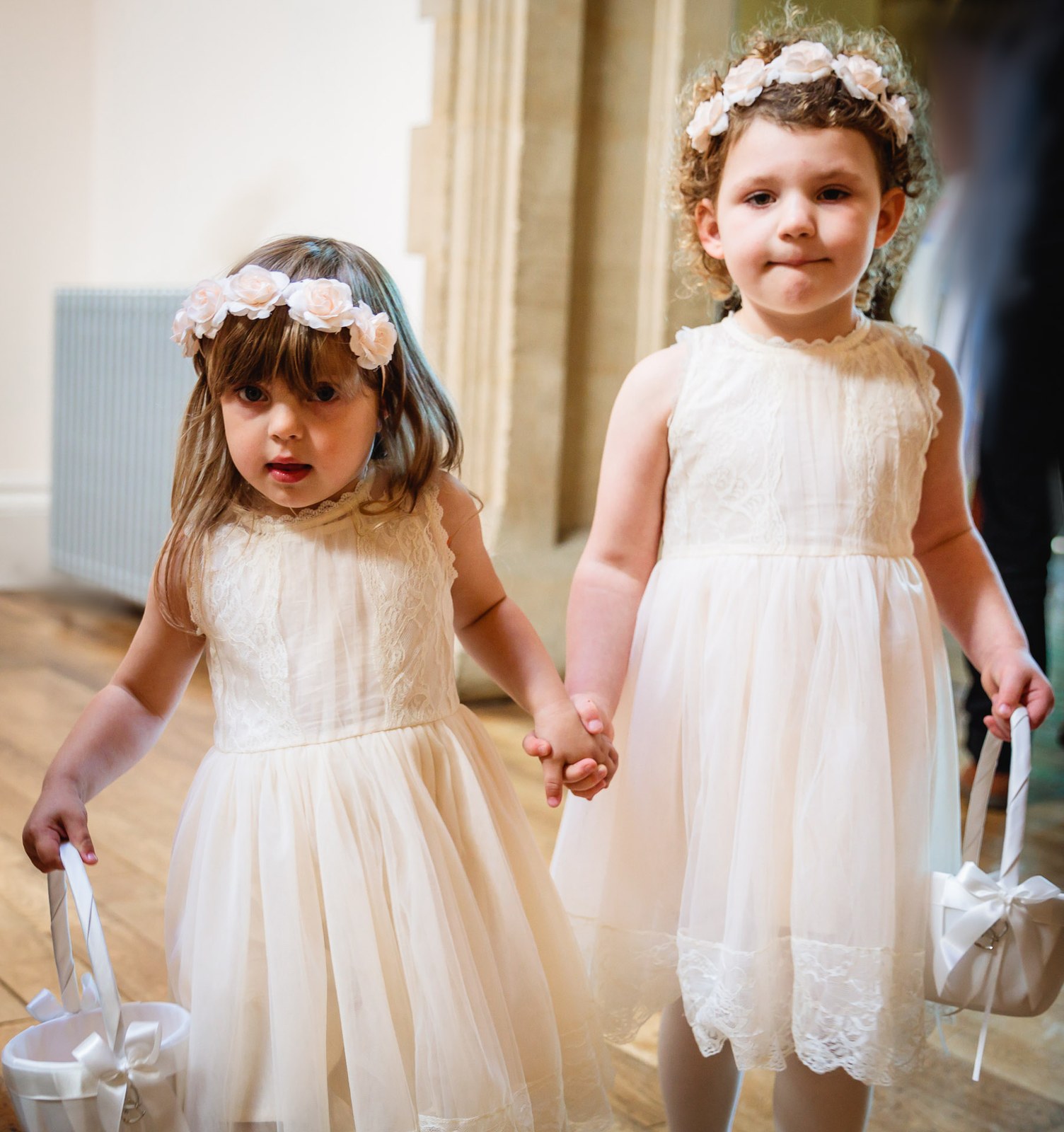 Two flower girls in cream with floral crowns hold hands, petal baskets in hand