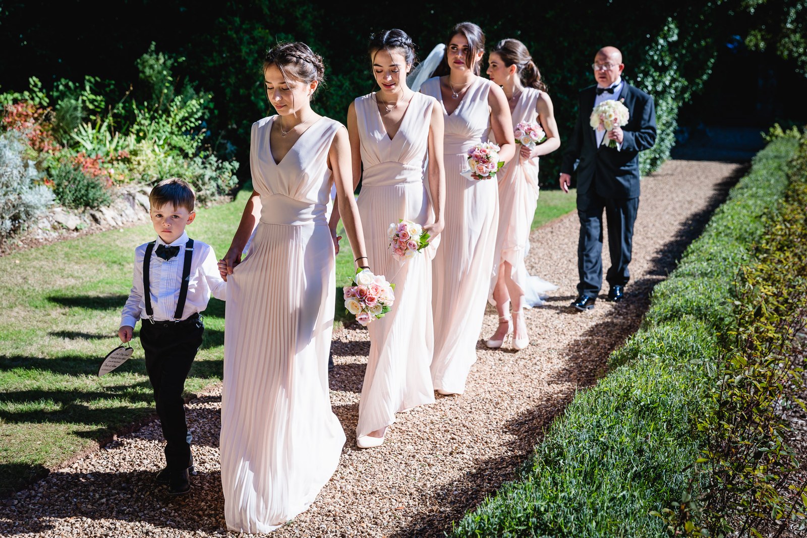Bridesmaids, a page boy and father walk a sunlit garden path at St Audries Park