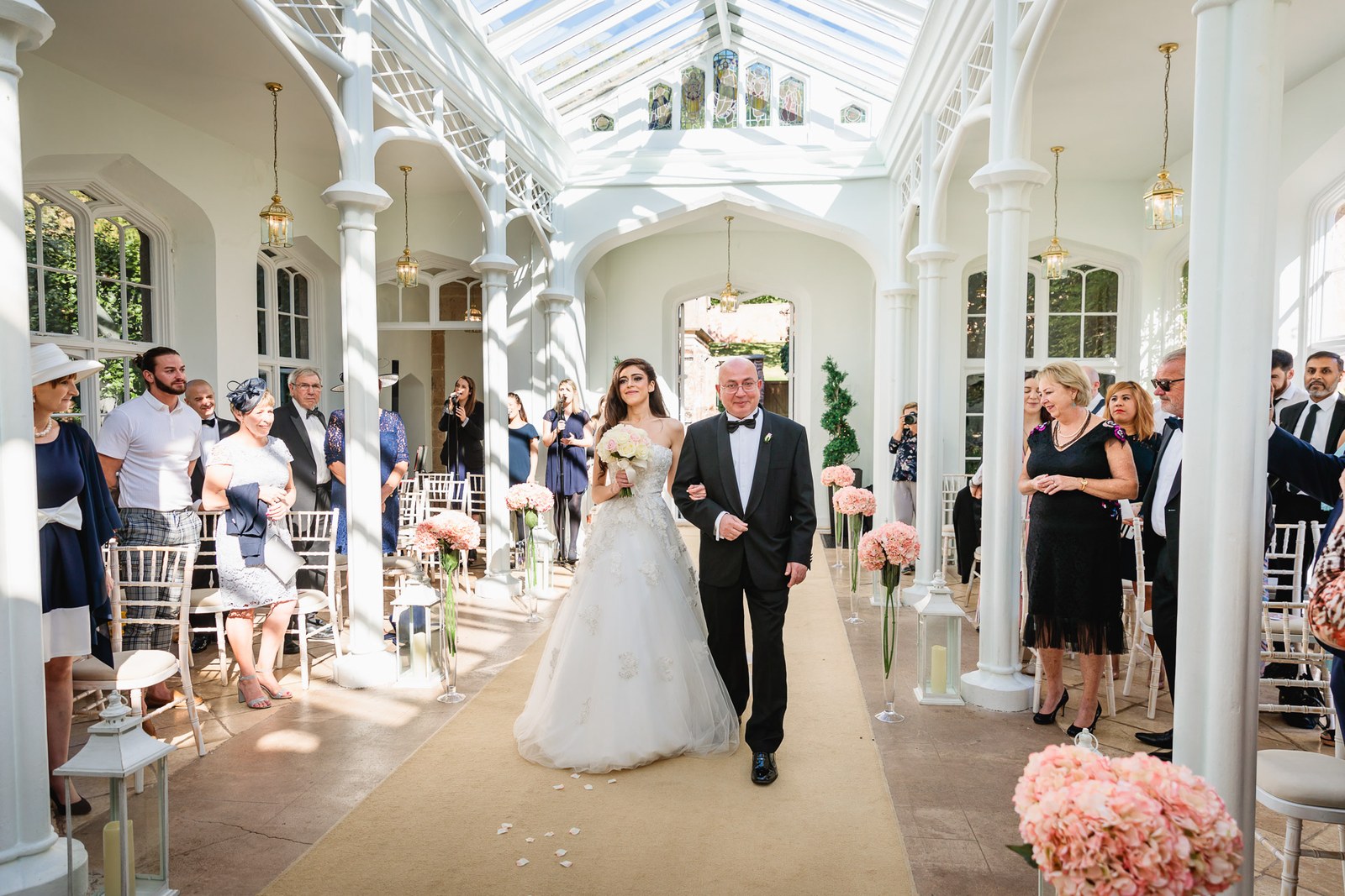 Bride on her father's arm walks the aisle through the glass orangery