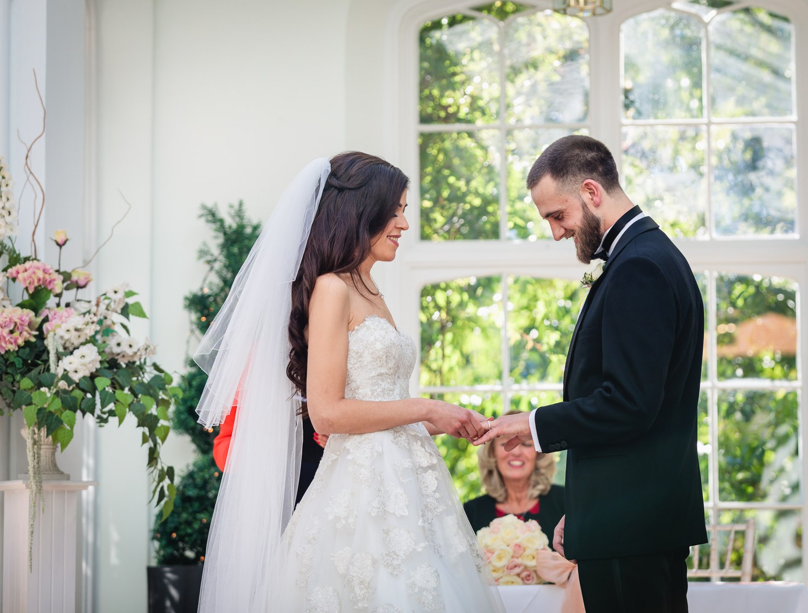 Groom places the ring on the bride's hand by the orangery windows