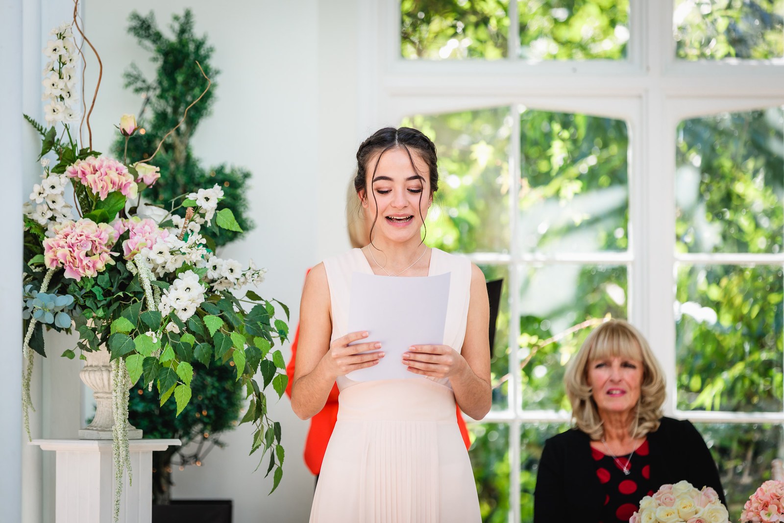 A young woman in blush gives a reading beside a tall floral arrangement