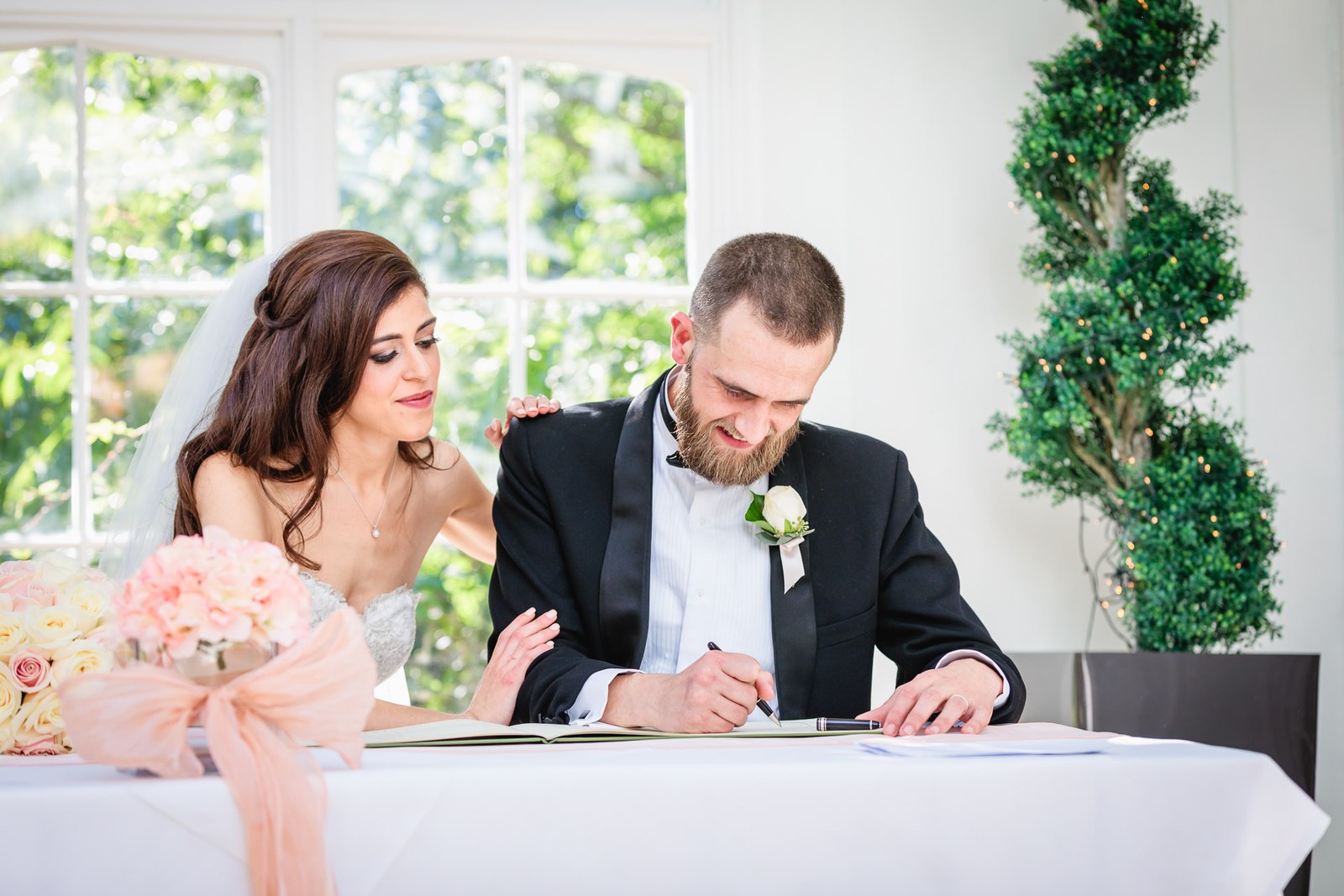 The groom signs the register as the bride leans in at the flower-dressed table