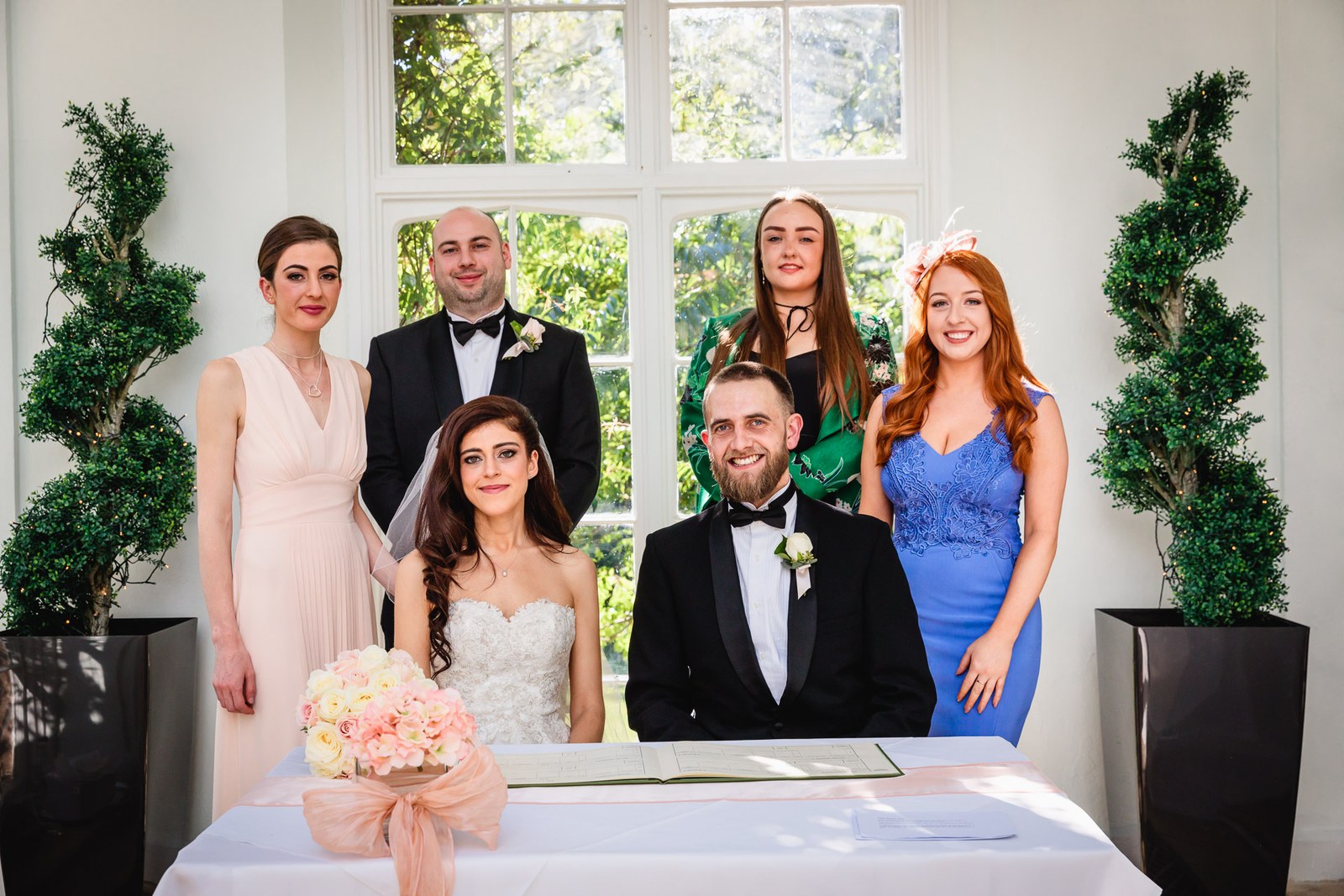 The wedding party gather behind the signing table between clipped topiary
