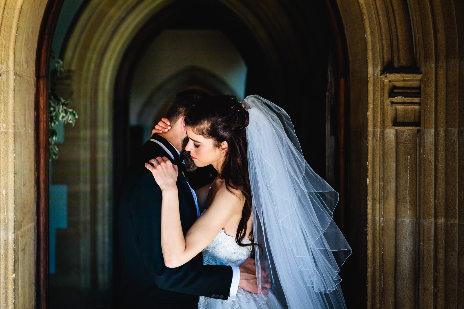The couple embrace within a shadowed Gothic archway, her veil catching light