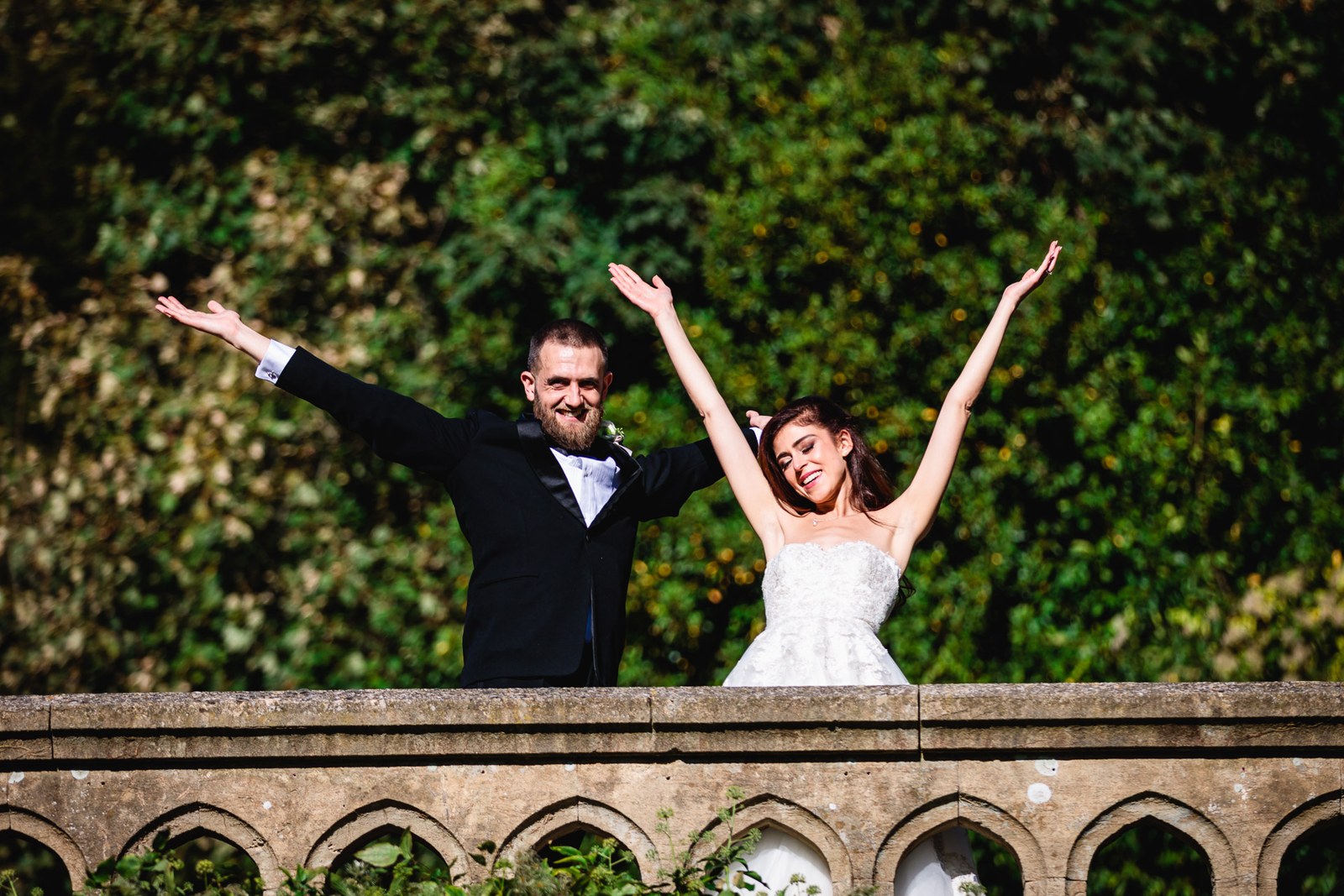 Bride and groom throw their arms wide on the stone balustrade above the gardens