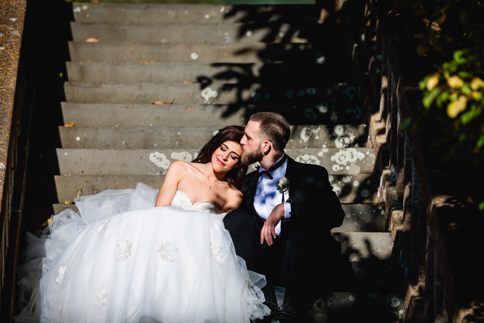 Groom kisses the bride's temple as they sit on the shaded stone garden steps