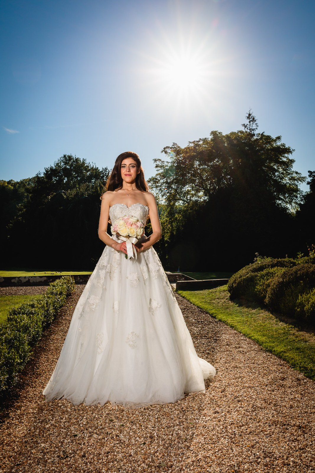 Bride on the garden path beneath a bright sunburst sky, bouquet in hand