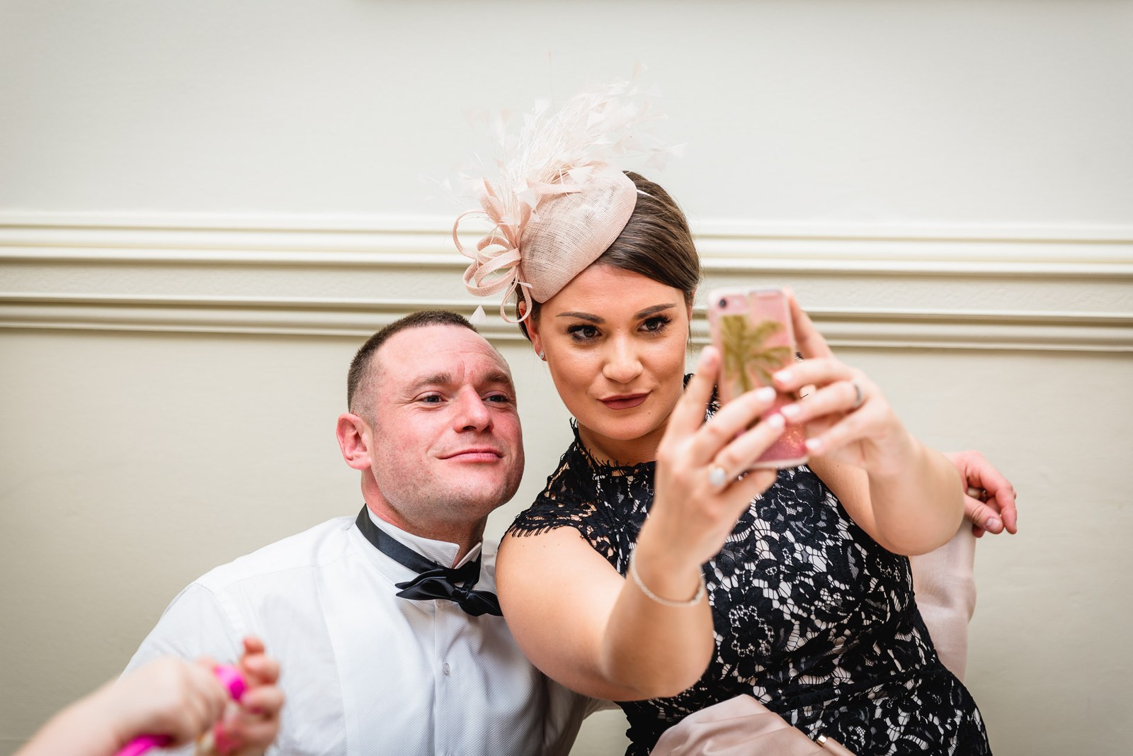 A guest in a feathered fascinator takes a selfie with a man in black tie