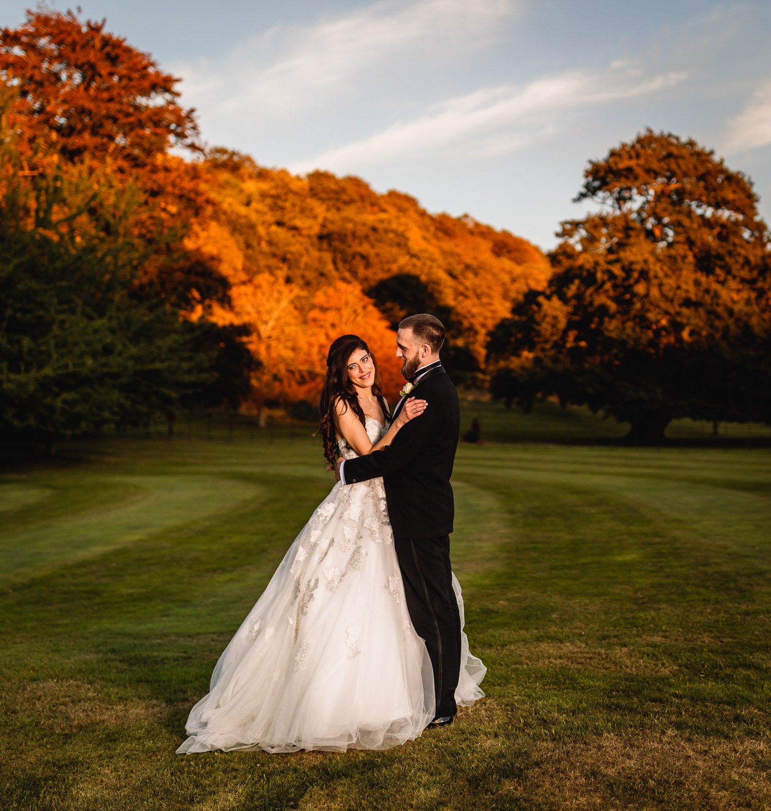The couple embrace on the lawn as autumn trees blaze gold behind them
