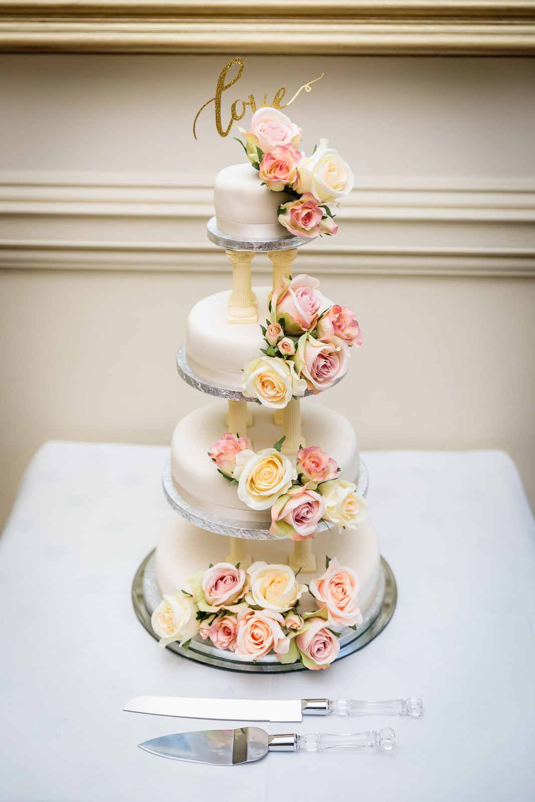 A four-tier white wedding cake dressed with roses and a gold 'love' topper