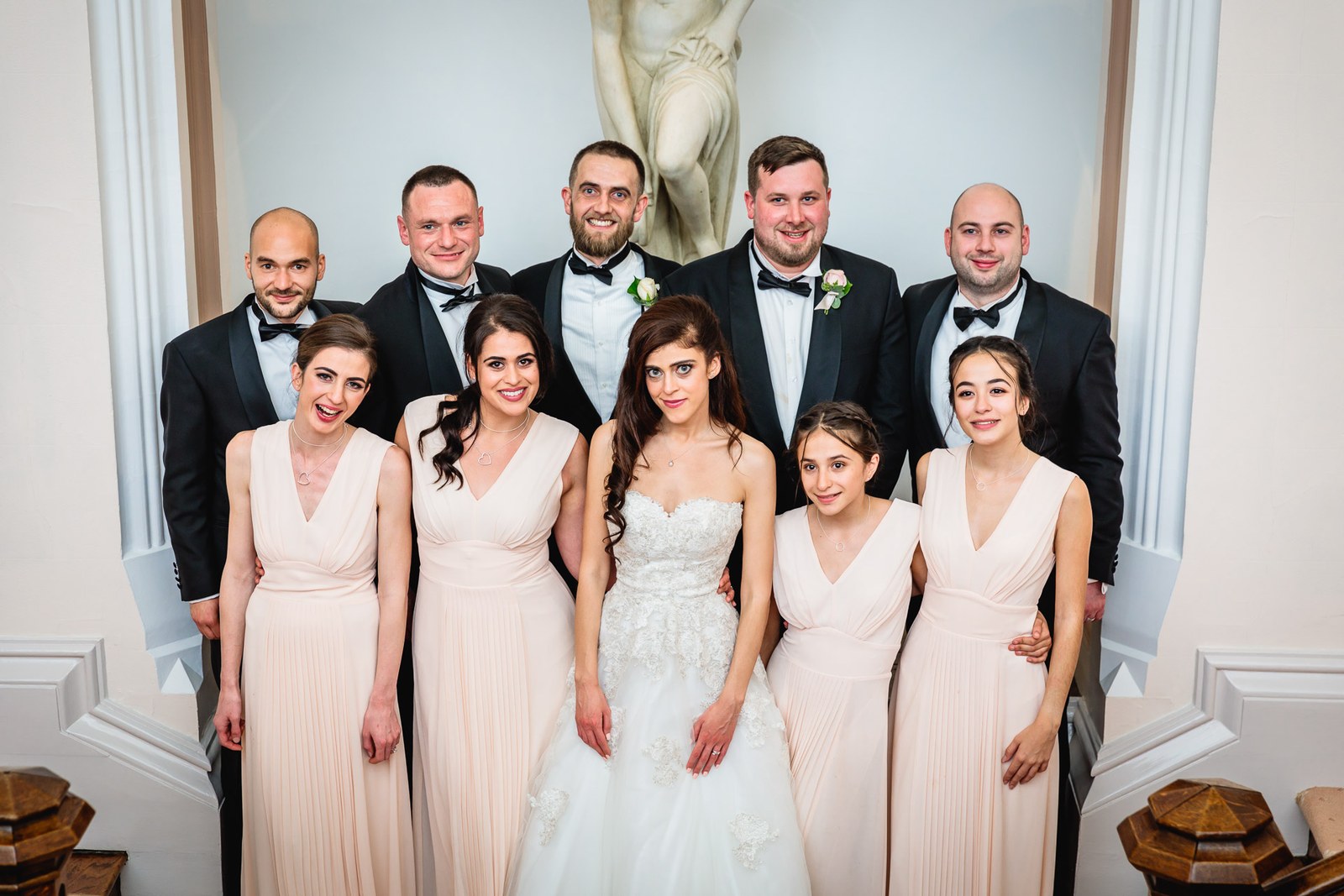 The wedding party in black tie and blush pose before a marble statue