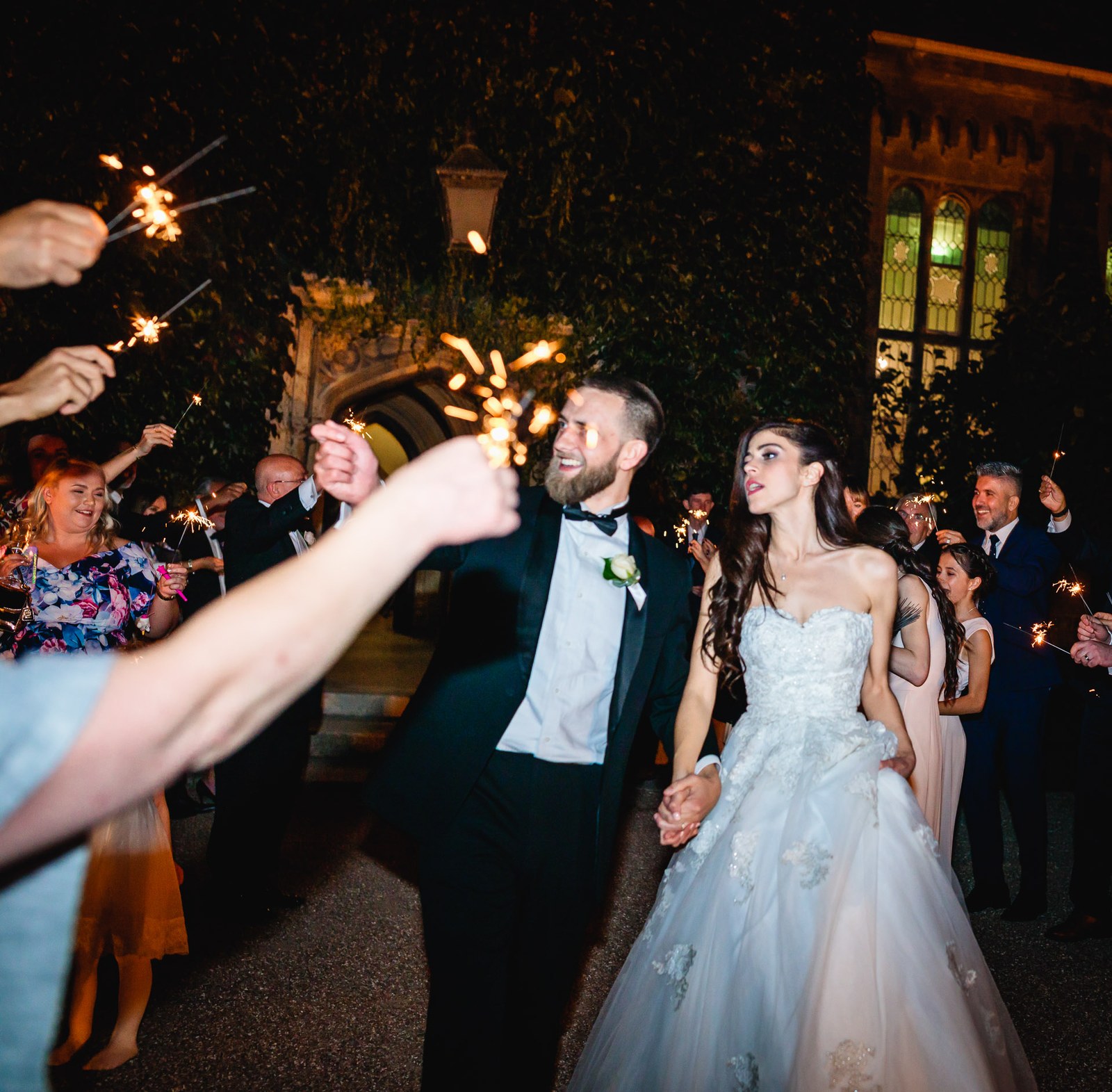 The couple walk a sparkler send-off before the floodlit Gothic manor at night