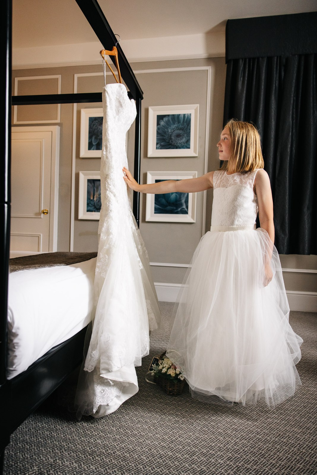 A young flower girl steadying the wedding dress on its hanger in the bedroom