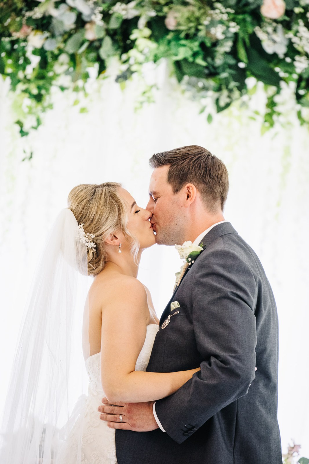 The couple kiss beneath the lush floral ceremony arch