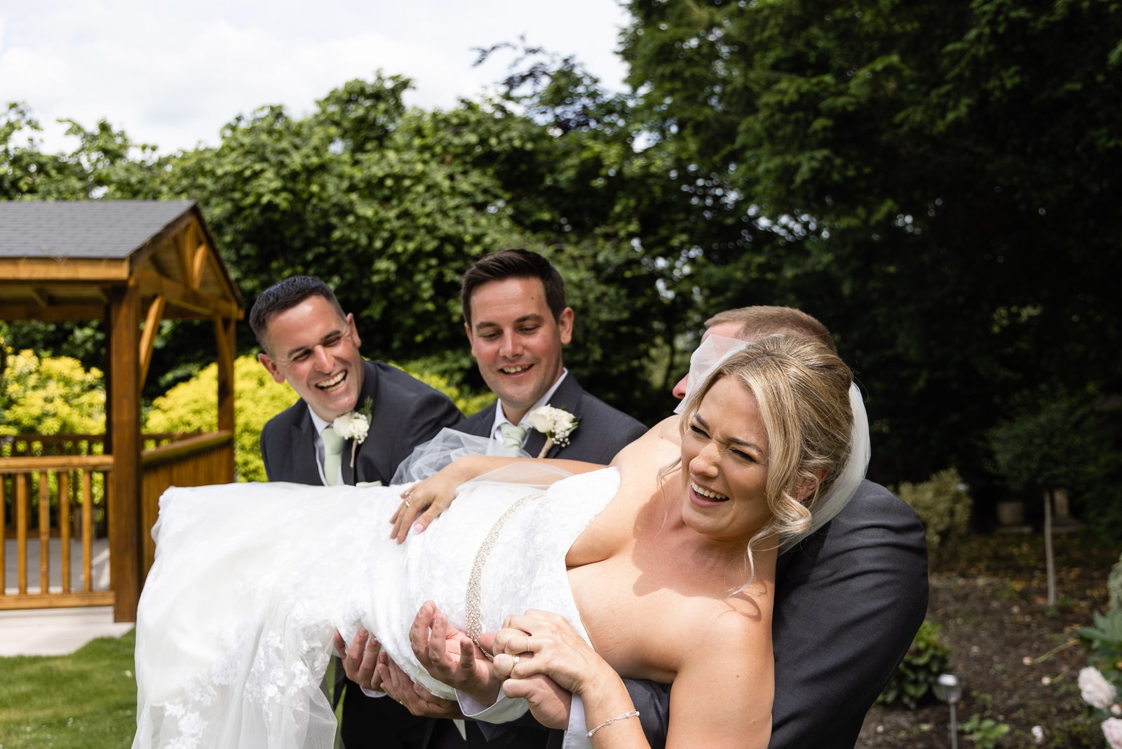 The groom and groomsmen cradling the laughing bride by the garden gazebo