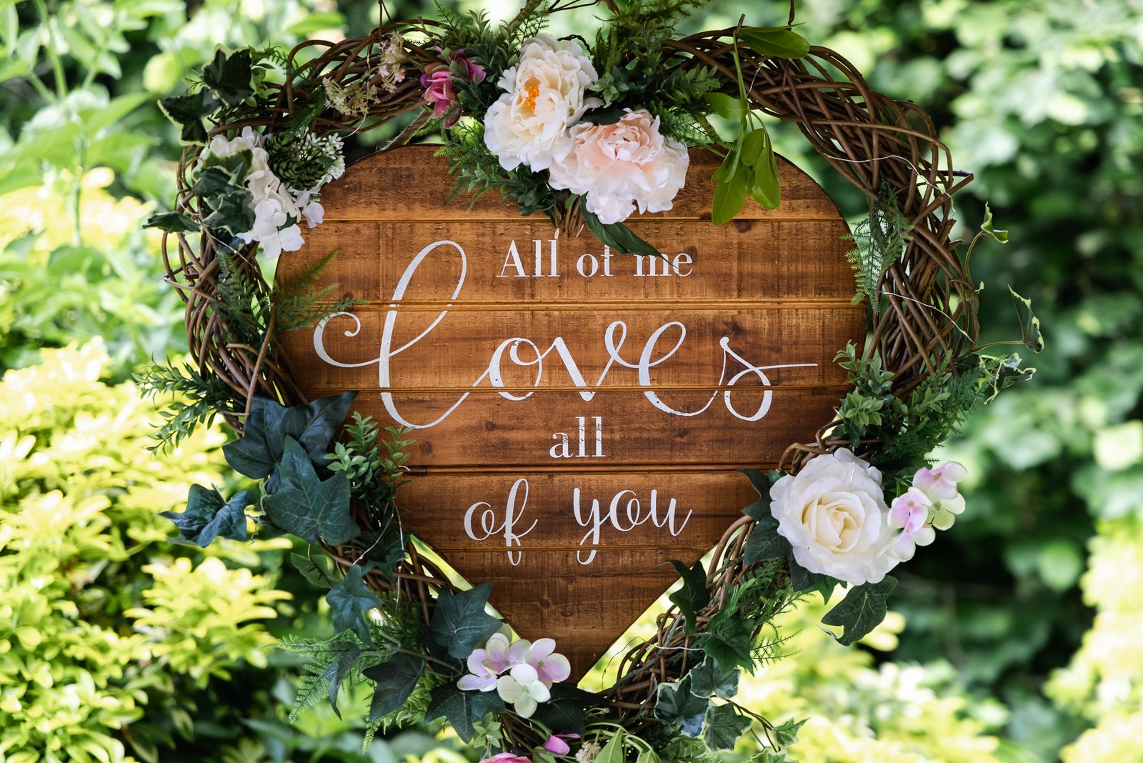 A rustic wooden heart sign reading 'all of me loves all of you' among foliage