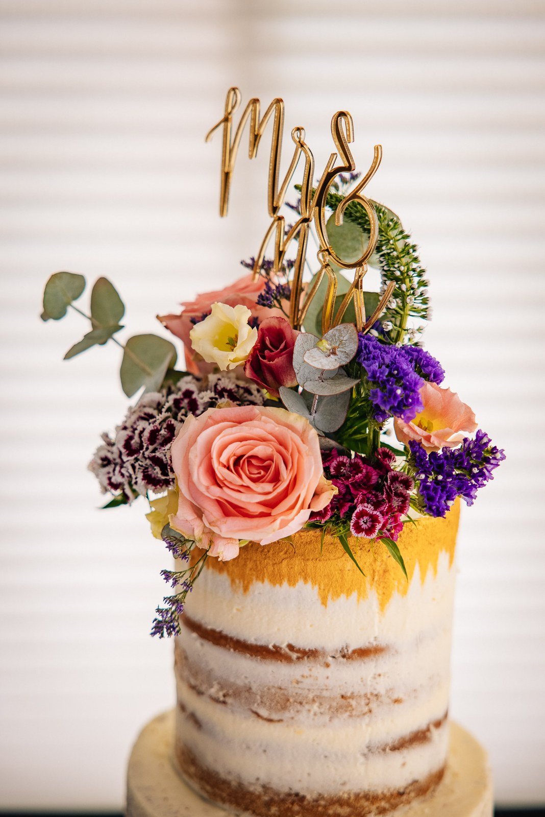 Semi-naked wedding cake topped with flowers and a gold script cake topper