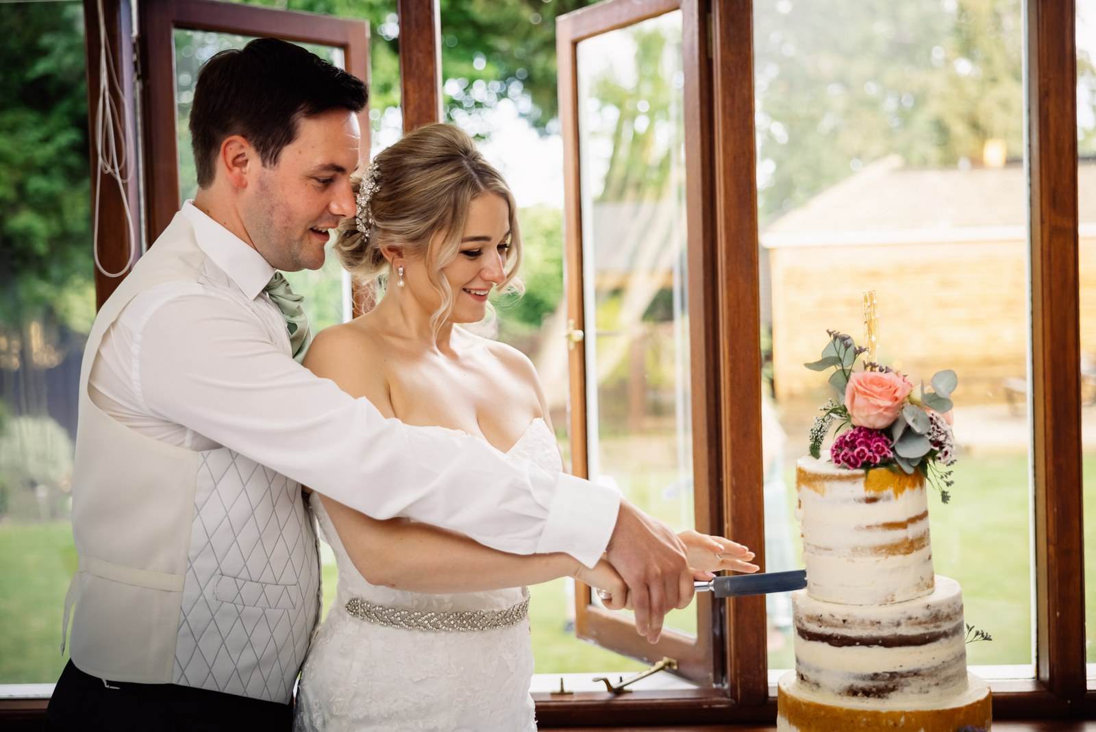 Bride and groom cutting their flower-topped cake by a window