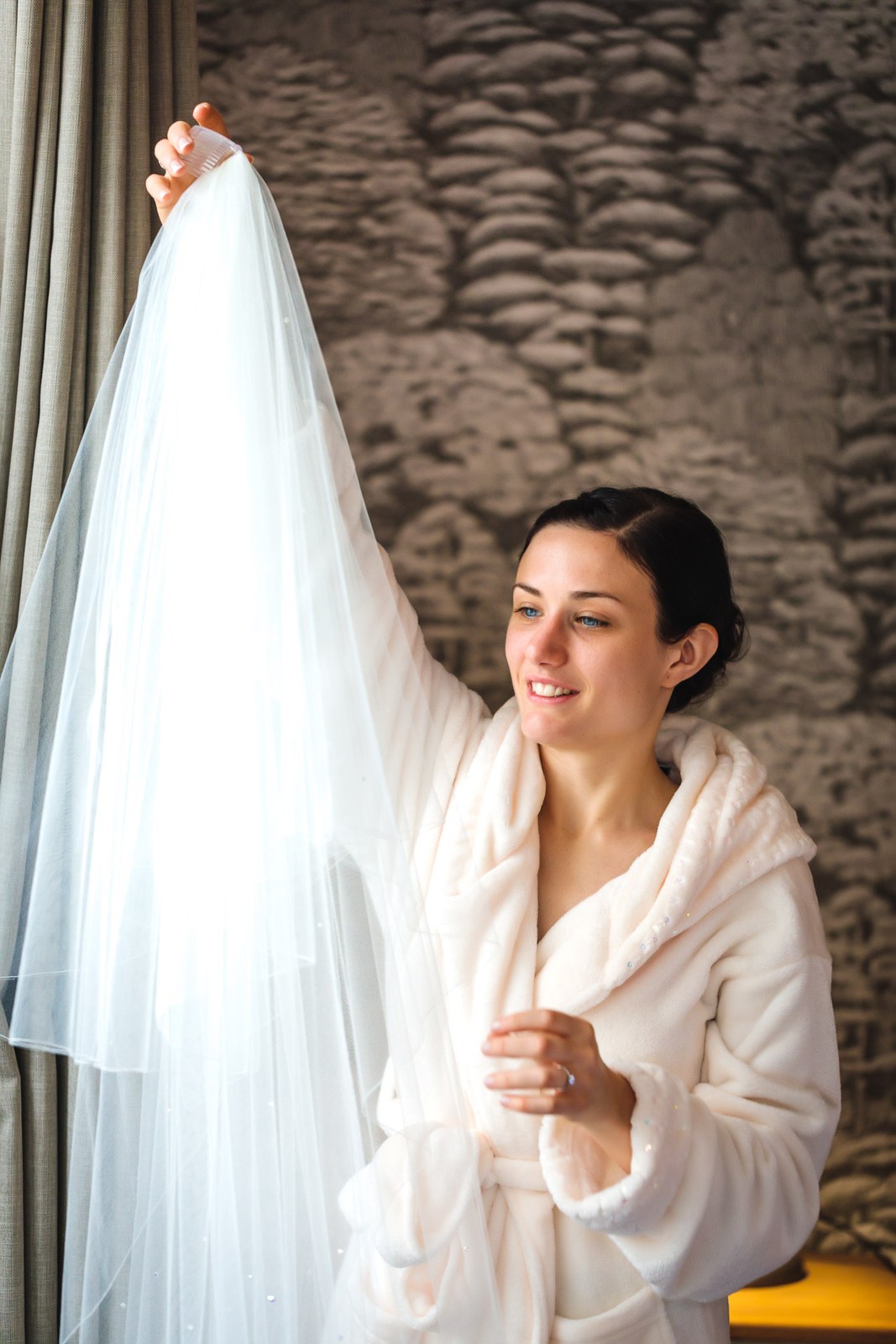Bride in a fluffy robe holds her veil aloft against an exposed stone wall