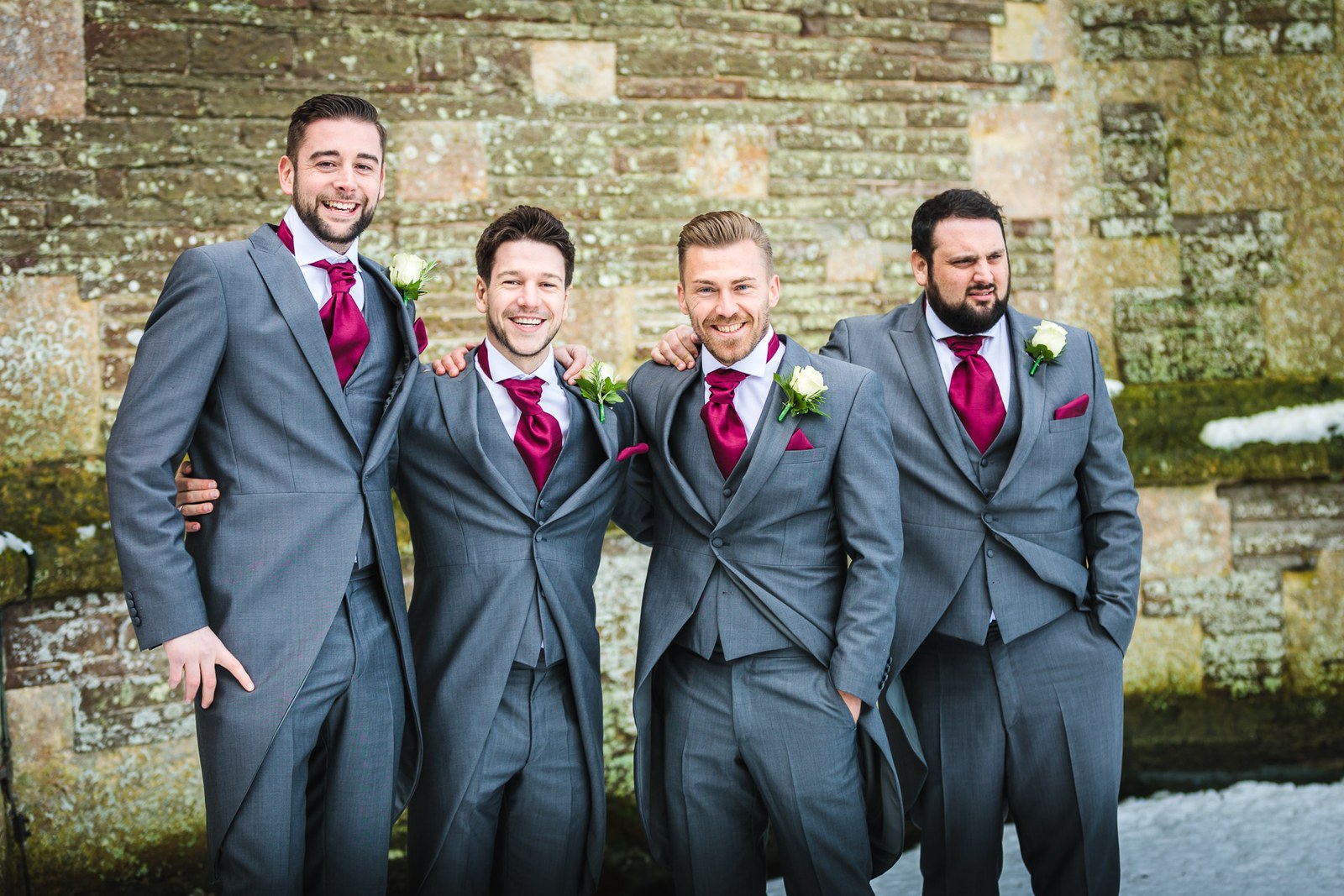 Groom and three groomsmen in grey morning suits and burgundy ties by a snow-dusted wall