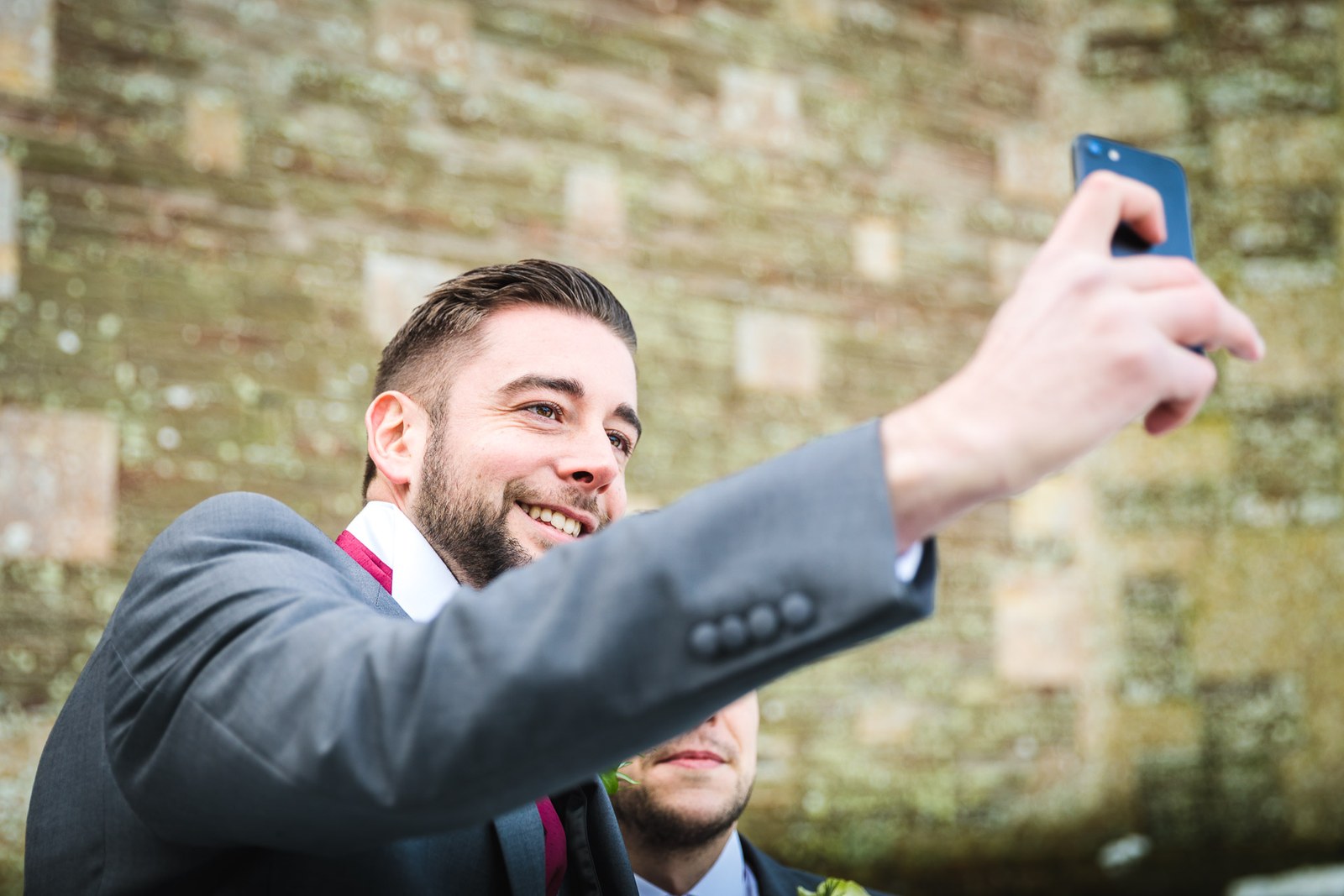 A groomsman grins as he takes a selfie against the old church stonework